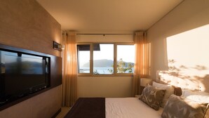 A hotel room with a bed, a TV mounted on the wall, a window with curtains, and a view of the sea. - Troia Residence Praia (Grandola)