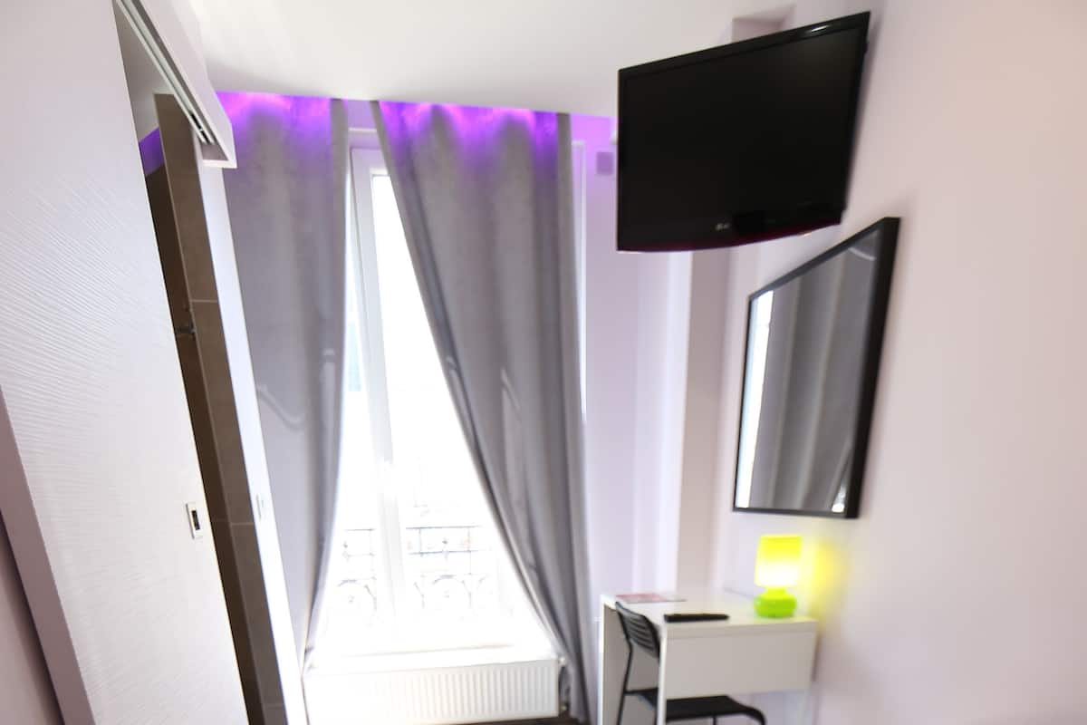 A hotel room with a bed, a desk, a chair, a TV mounted on the wall, and a window with curtains.