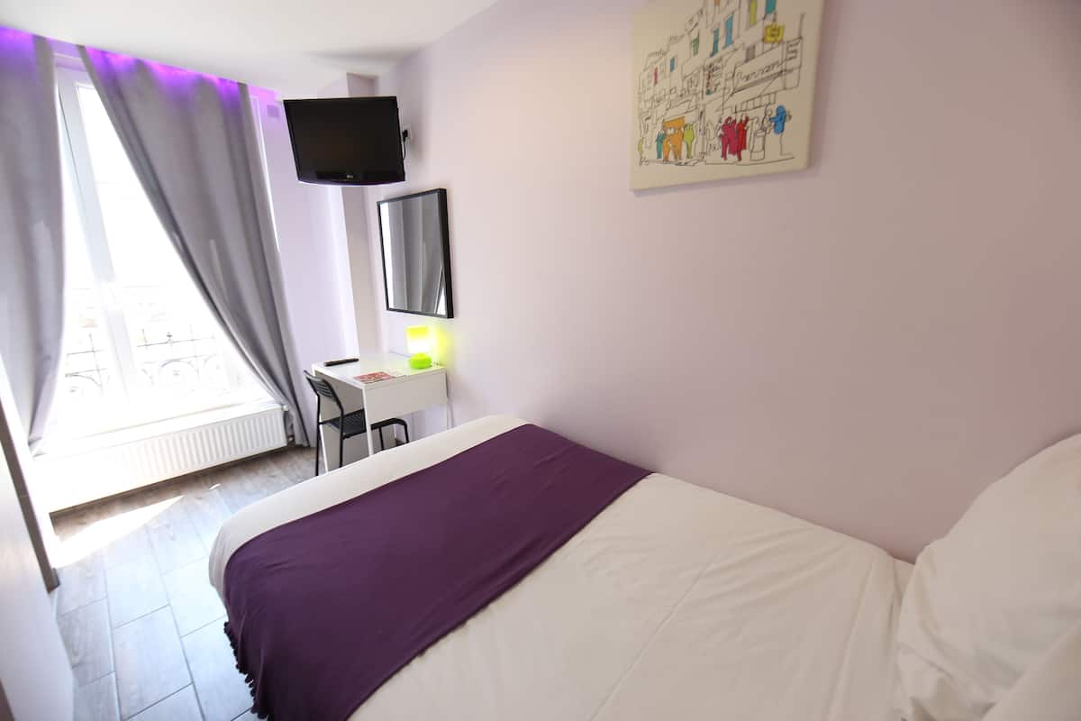 A bedroom with a bed, a desk, a chair, a flat-screen TV, and a window with curtains.