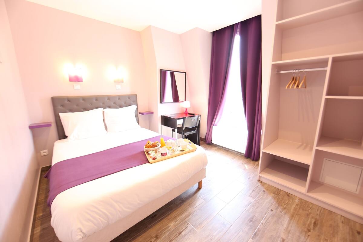 A hotel room with a large bed, a bedside table with a lamp, a wardrobe, and a tray with breakfast items.