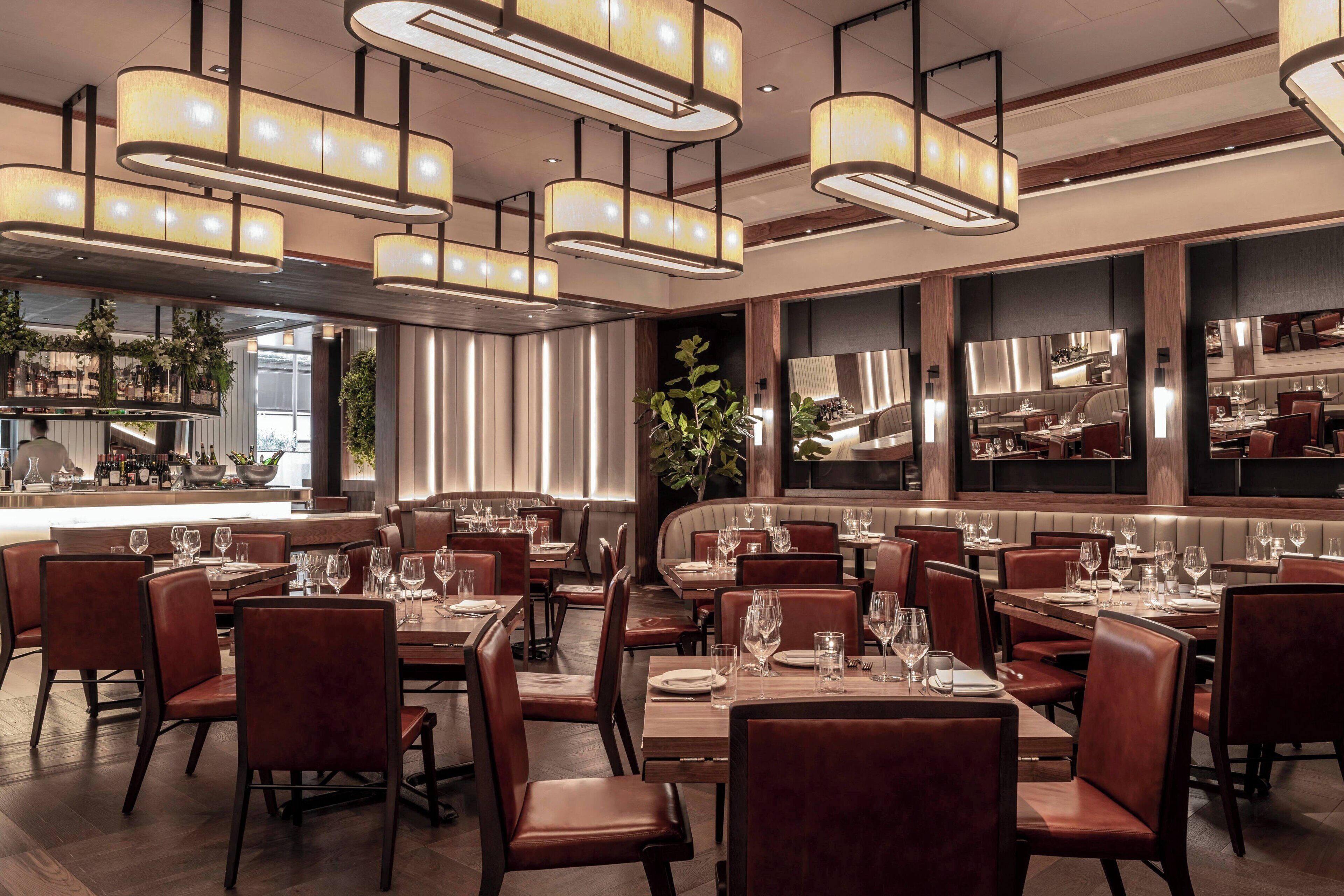 A restaurant with multiple dining tables and chairs, a bar area, and a kitchen visible through a glass partition.