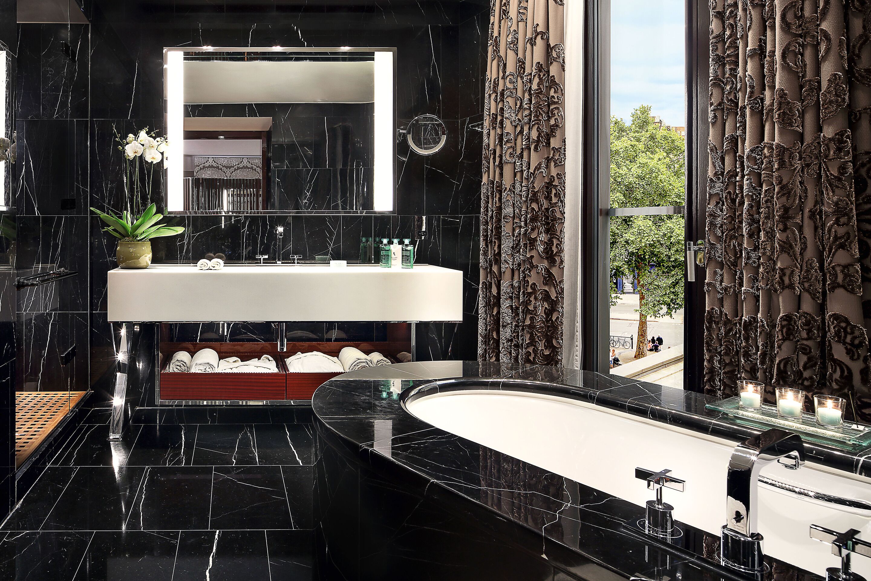 Une salle de bain moderne avec un grand miroir, des plans de travail en marbre noir, une baignoire sur pieds et une vue sur l’extérieur.