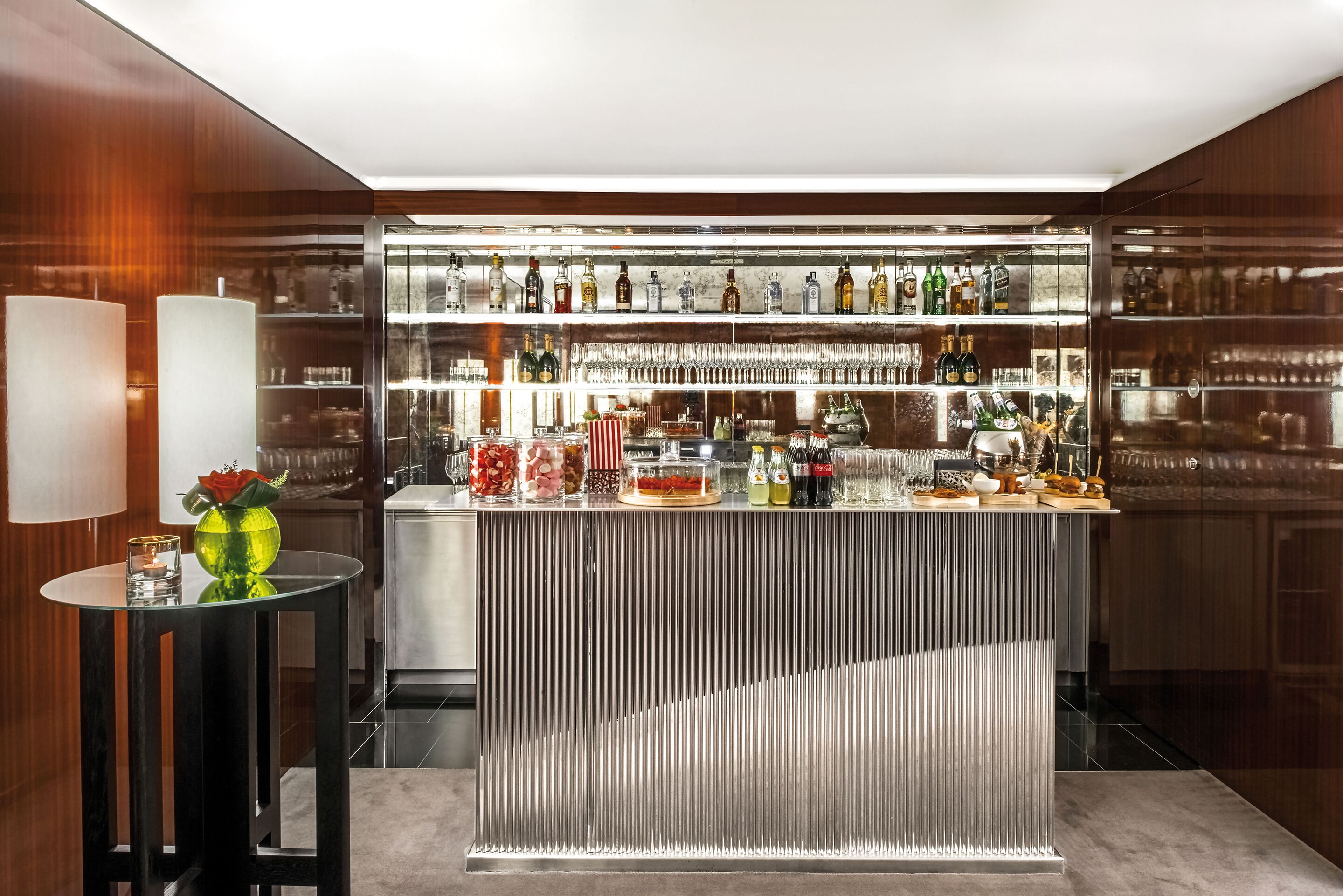 Un coin bar avec un présentoir en verre rempli de bouteilles, une table ronde ornée d’un vase et d’un objet décoratif.