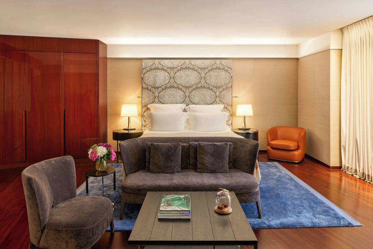 A hotel room with a large bed, two bedside lamps, a sofa, a coffee table with books, a chair, and a wardrobe.