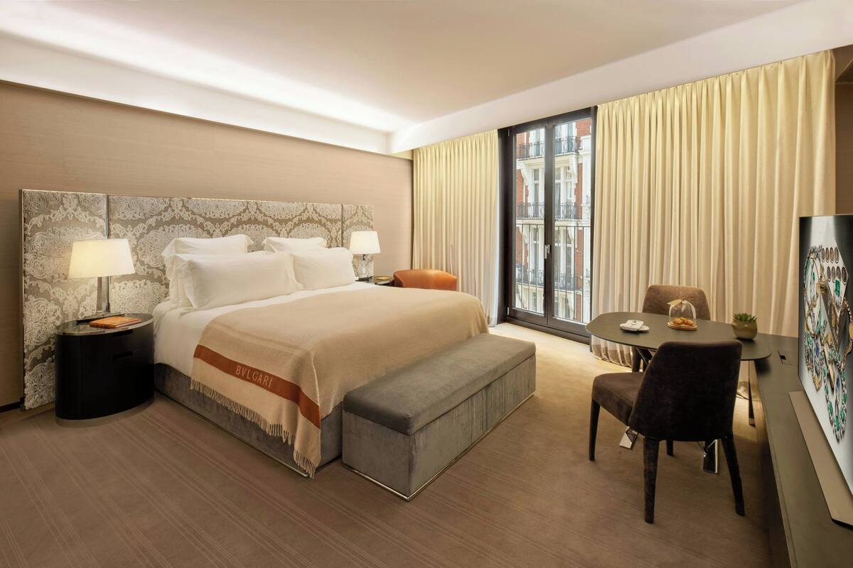 A hotel room with a large bed, a bedside table with a lamp, a small table with a chair, and a large window with curtains.