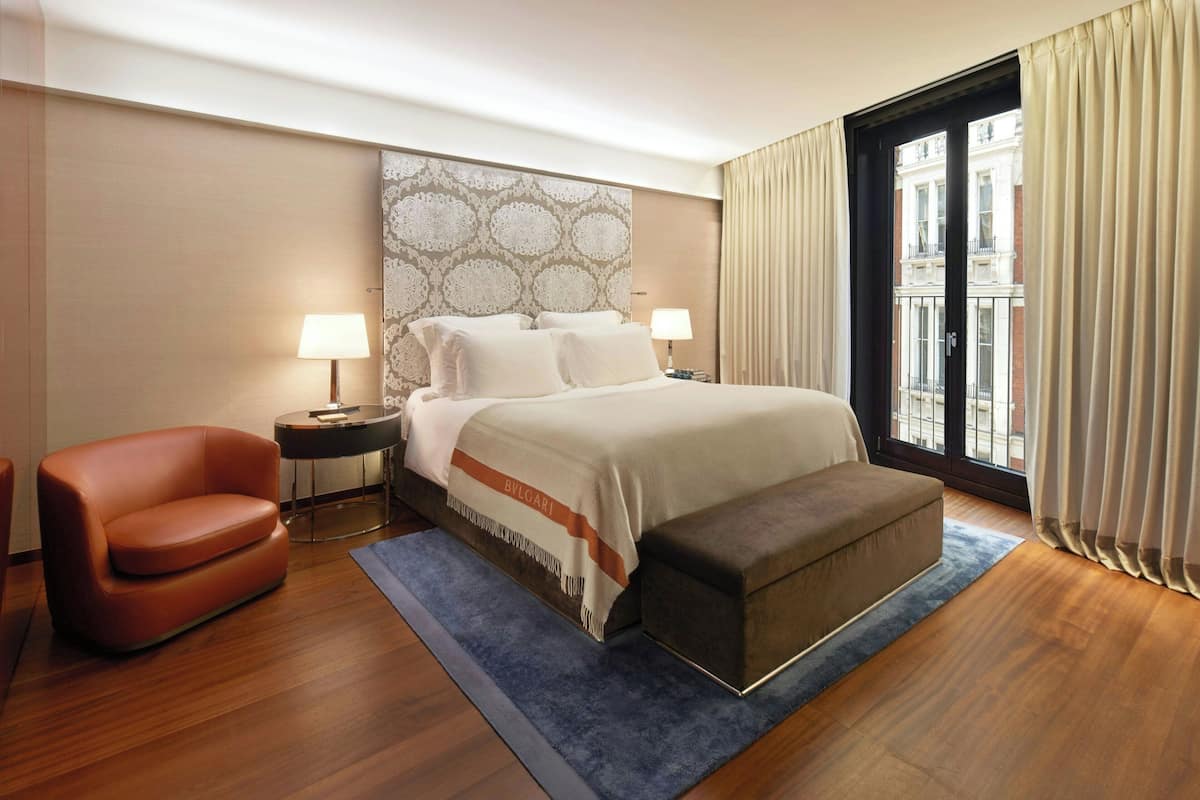A hotel room with a large bed, two bedside tables with lamps, a sofa, and a view of buildings through a window.