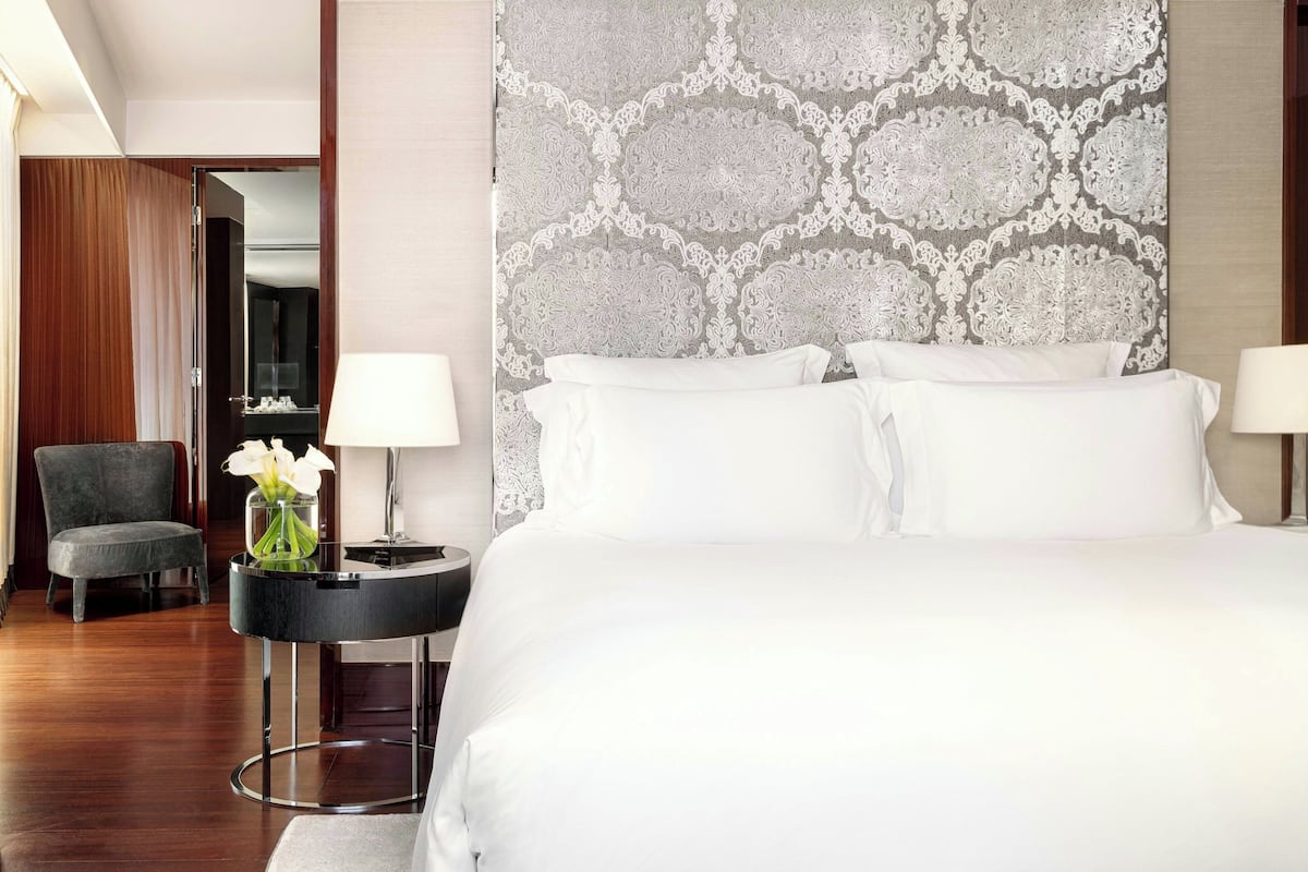 A hotel room with a large bed, two bedside lamps, a round side table with a vase of flowers, a chair, and a patterned headboard.