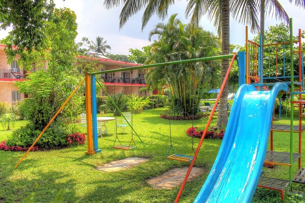 A playground with swings and a slide, surrounded by lush greenery and a building in the background. - Hotel Posada Koniapan (Catemaco)
