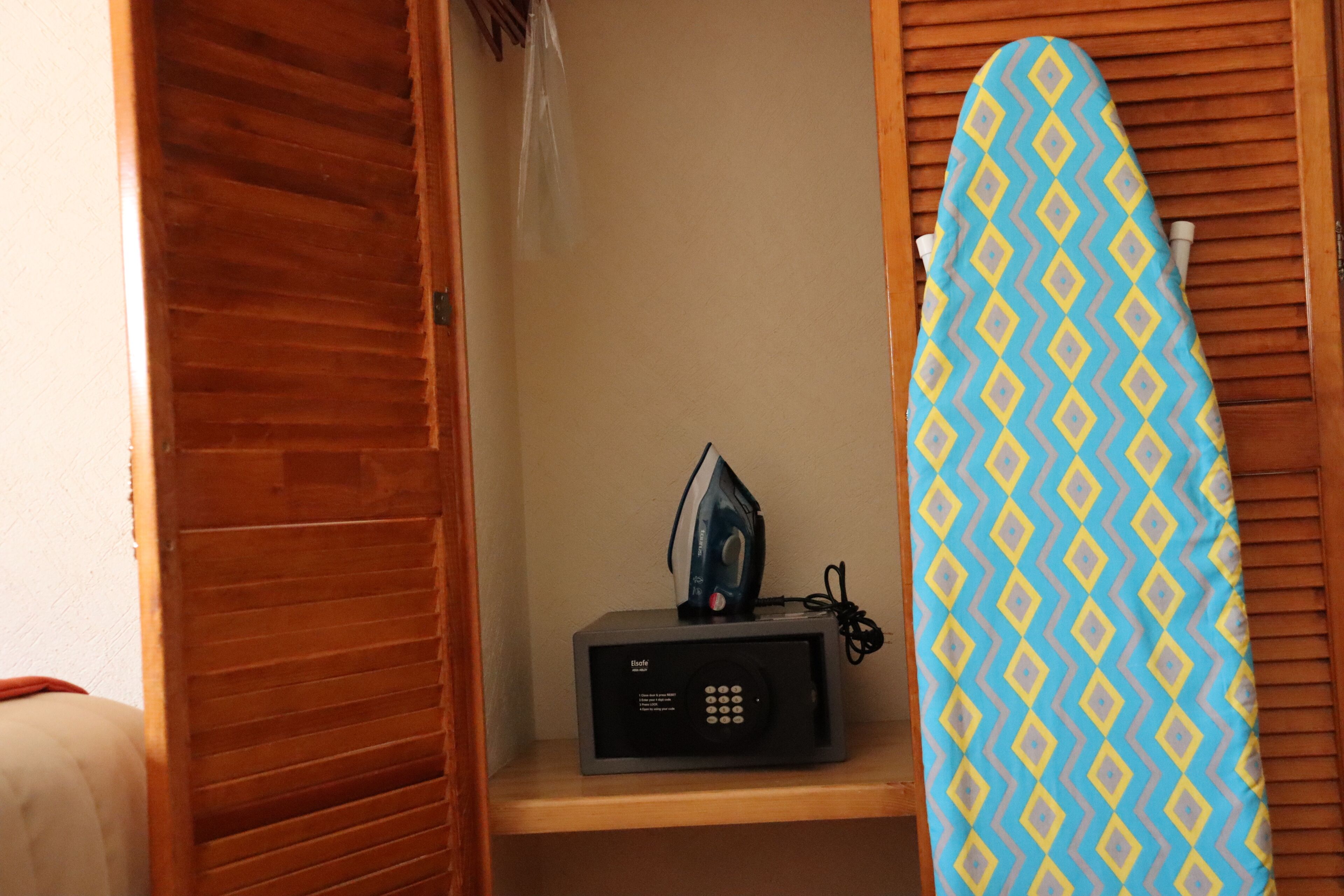 A small room with a wooden door, a shelf with an iron and a microwave, and a patterned surfboard leaning against the wall.