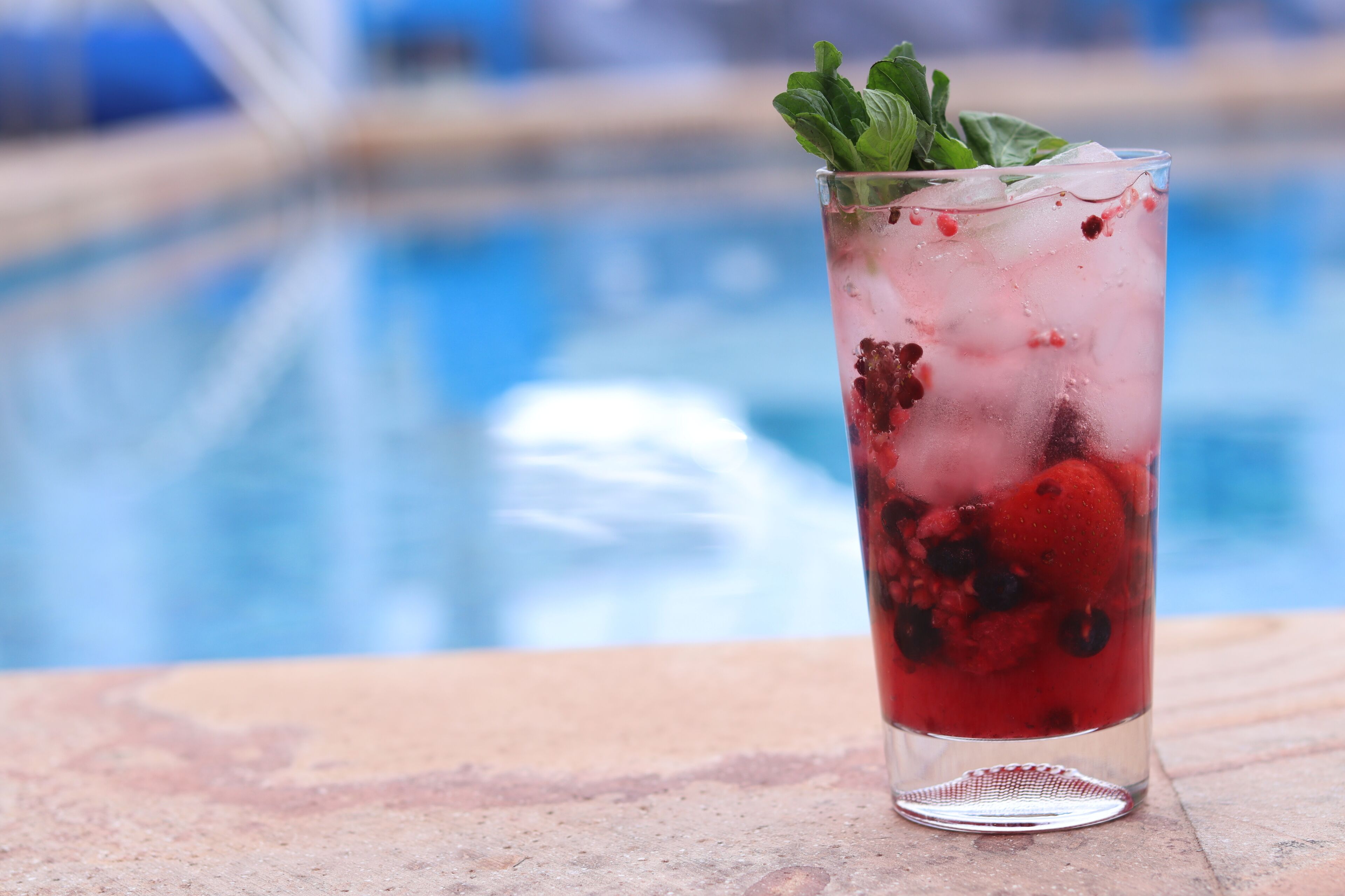 A tall glass with ice and berries, placed on a stone surface near a pool.