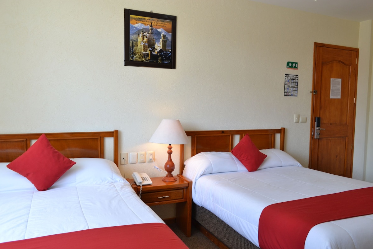 Habitación de hotel con dos camas, cada una con ropa de cama roja y blanca, cabeceros de madera, mesitas de noche con lámpara, teléfono y un cartel en la pared.