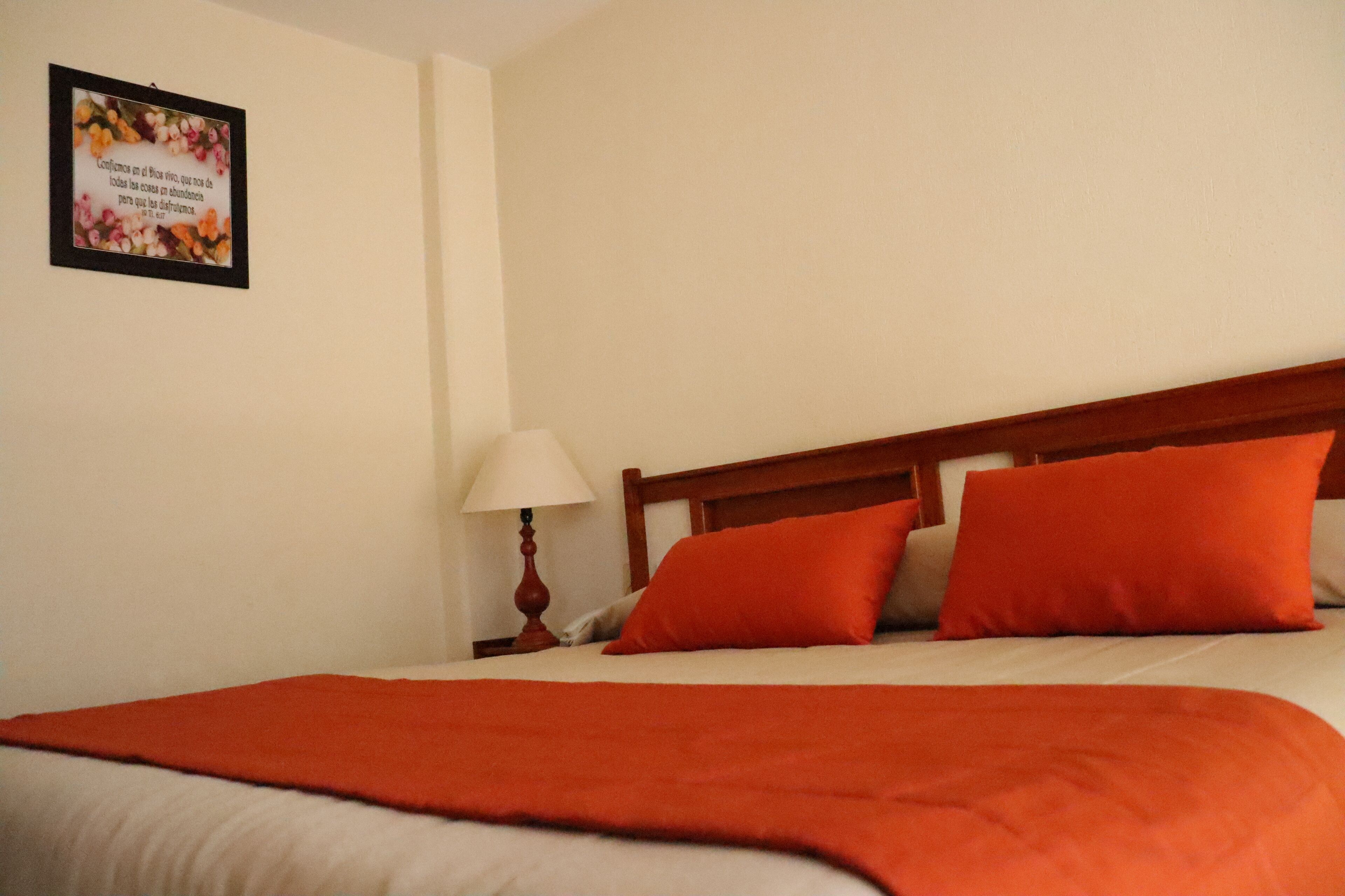 A neatly made bed with an orange blanket and pillows, a wooden headboard, a bedside lamp, and a framed quote on the wall.