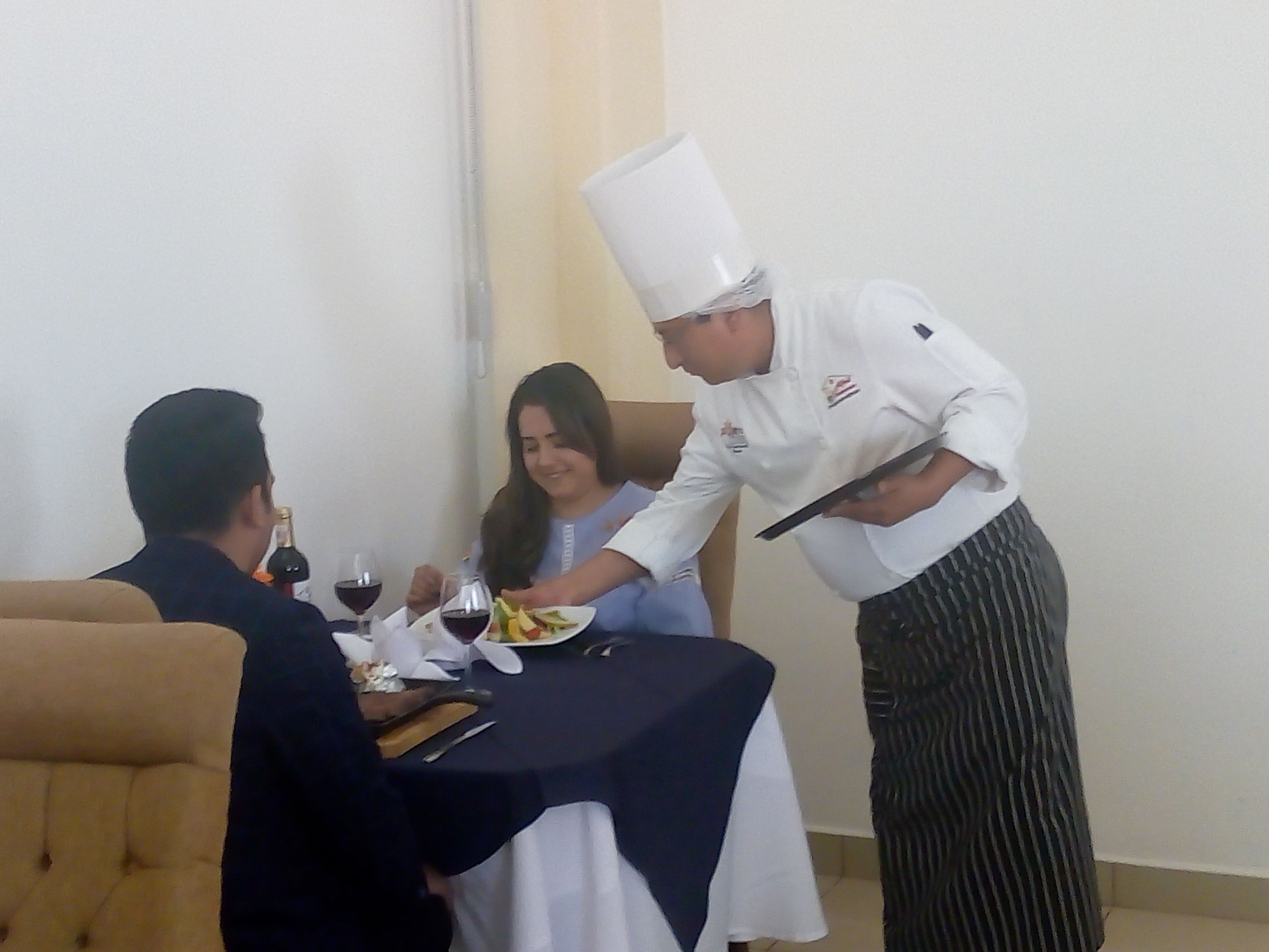 A dining area with a table set for two, a waiter serving a meal, and a bottle of wine on the table.