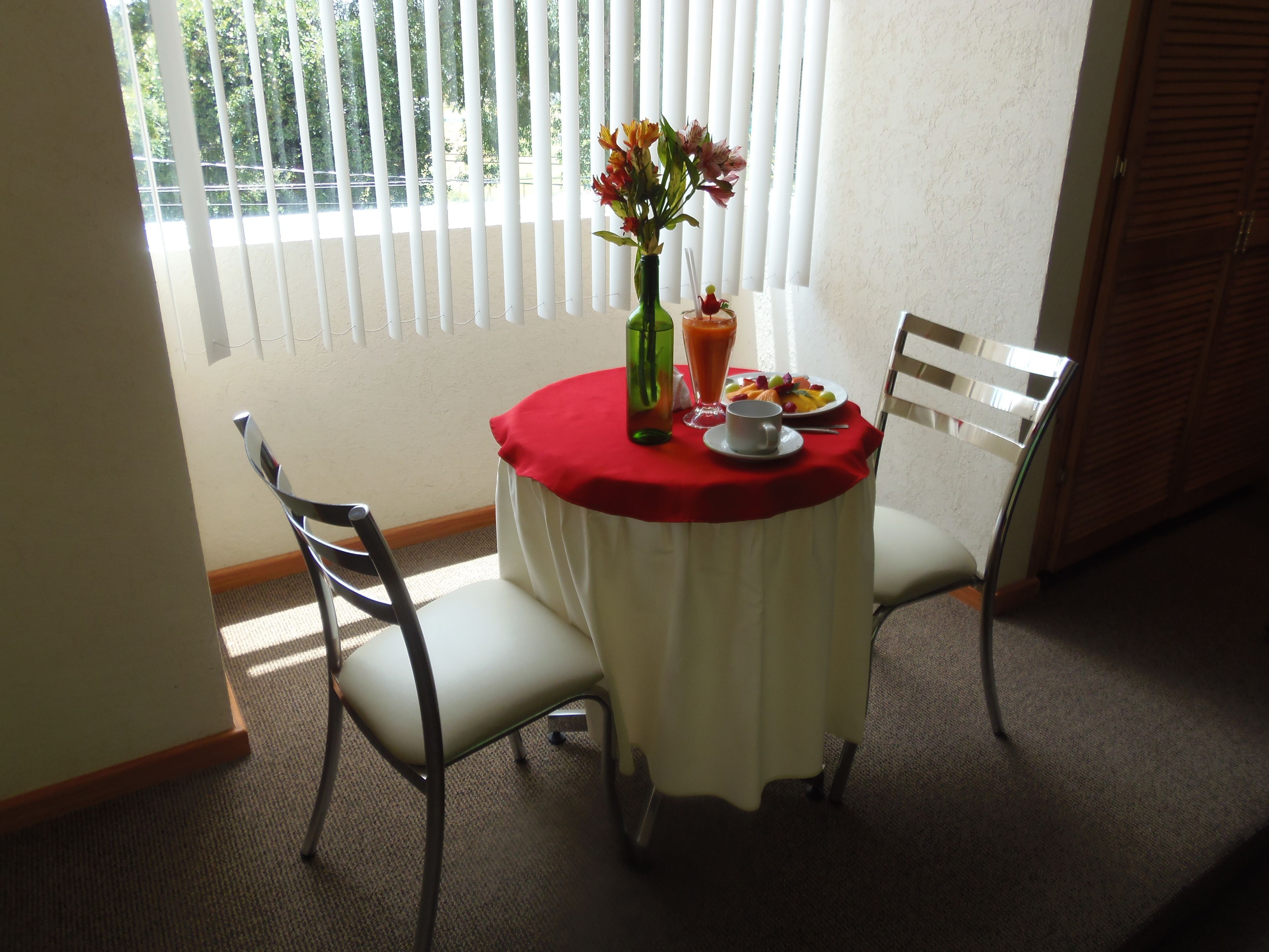 A dining table with a red tablecloth, two chairs, a vase with flowers, a glass of juice, and a plate of fruit.