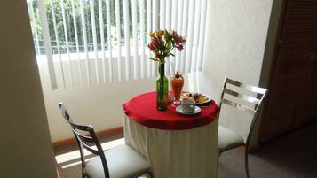 A dining table with a red tablecloth, two chairs, a vase with flowers, a glass of juice, and a plate of fruit.