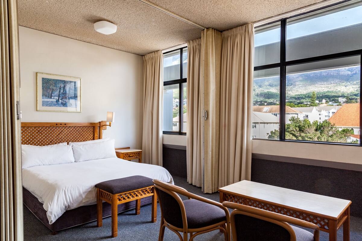 Ett hotellrum med en säng, två stolar, ett litet bord, ett nattduksbord och ett fönster med gardiner.