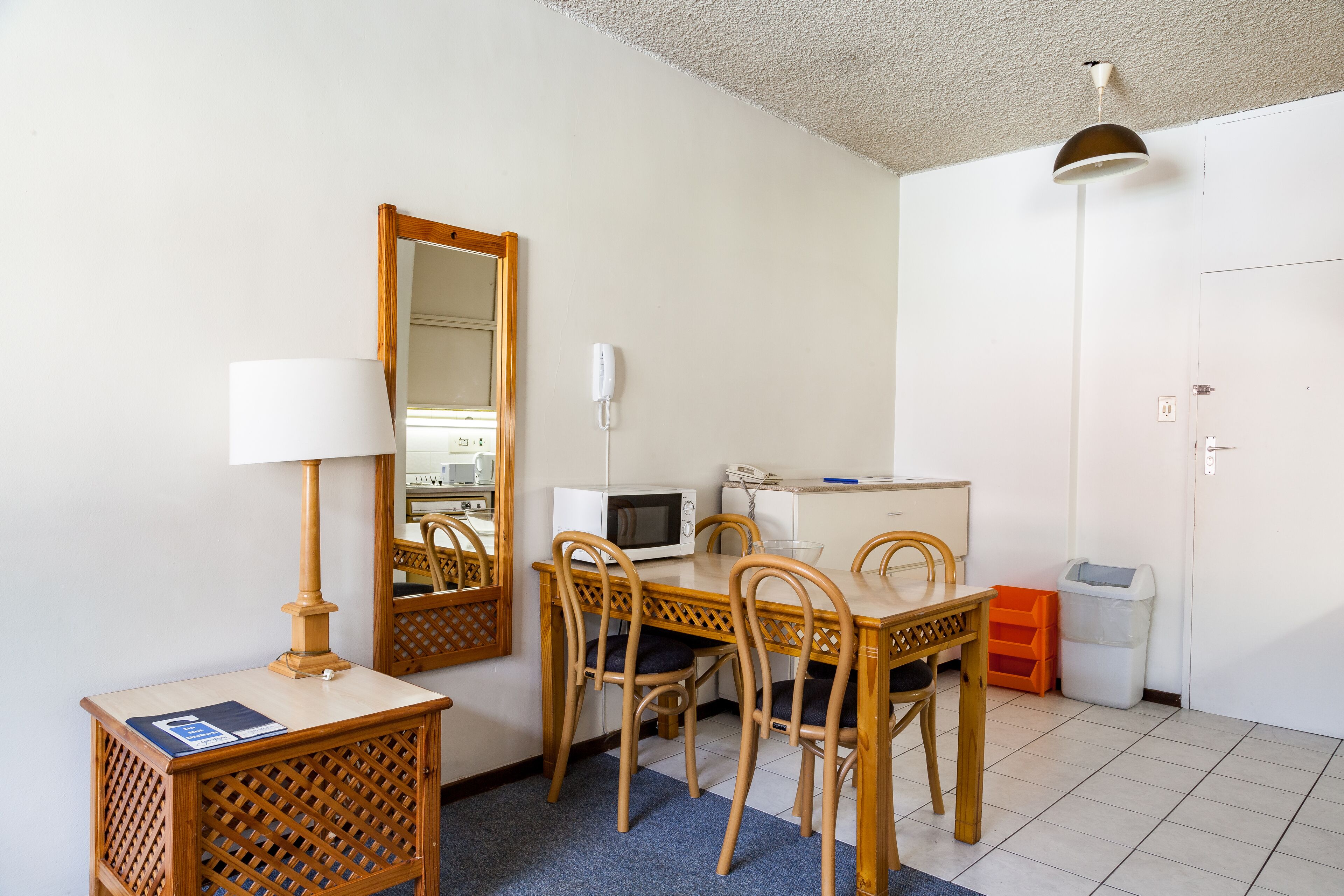 Habitación compacta con mesa de comedor, sillas, microondas, lámpara, espejo y cubo de basura.
