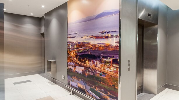 A modern elevator with a large panoramic view of a coastal cityscape, including a harbor and a bridge. - Gardens Centre Holiday Apartments (Cape Town)