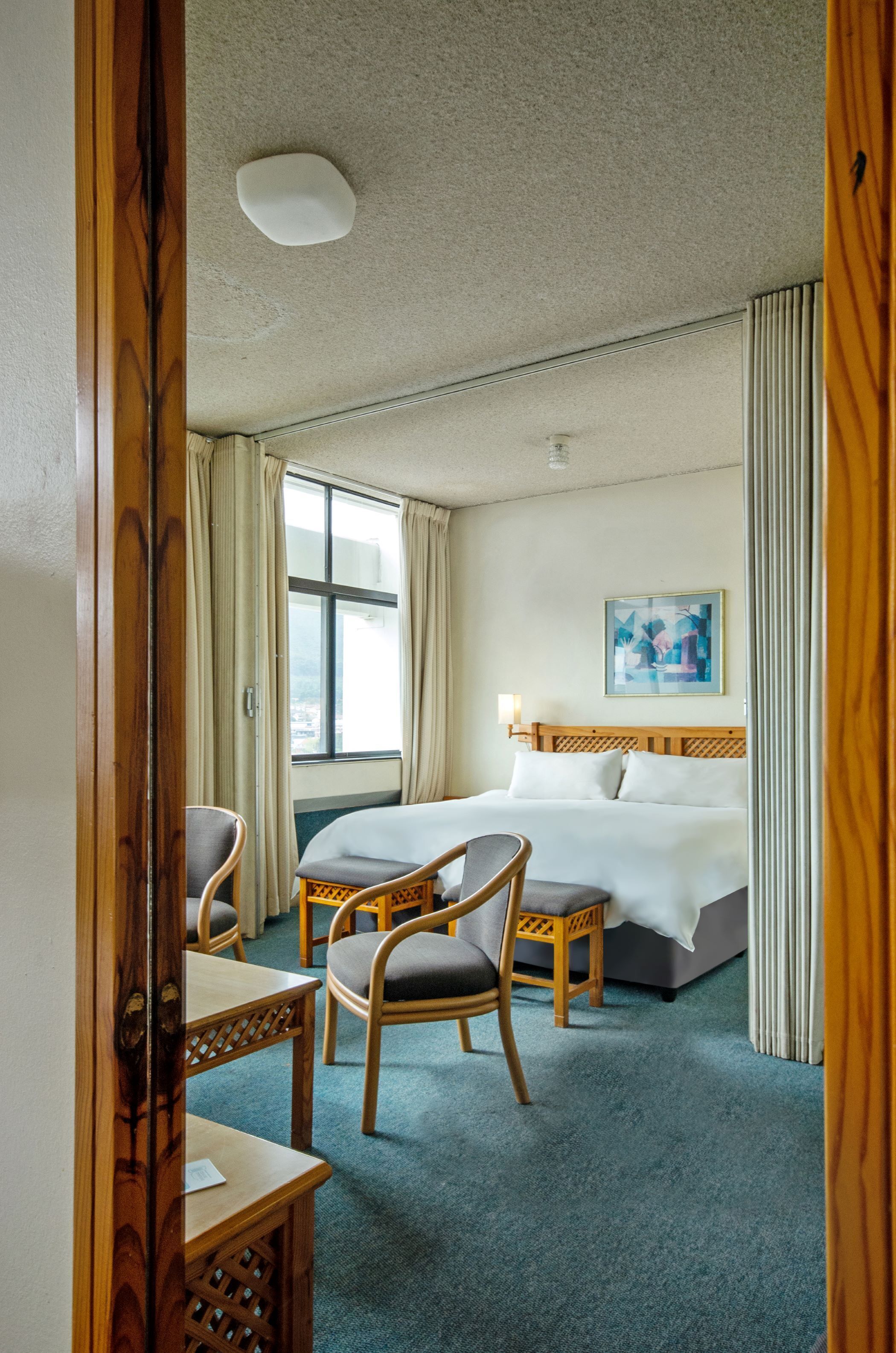 Habitación de hotel con una cama, dos sillas, una mesita, una ventana con cortinas y un cuadro en la pared.