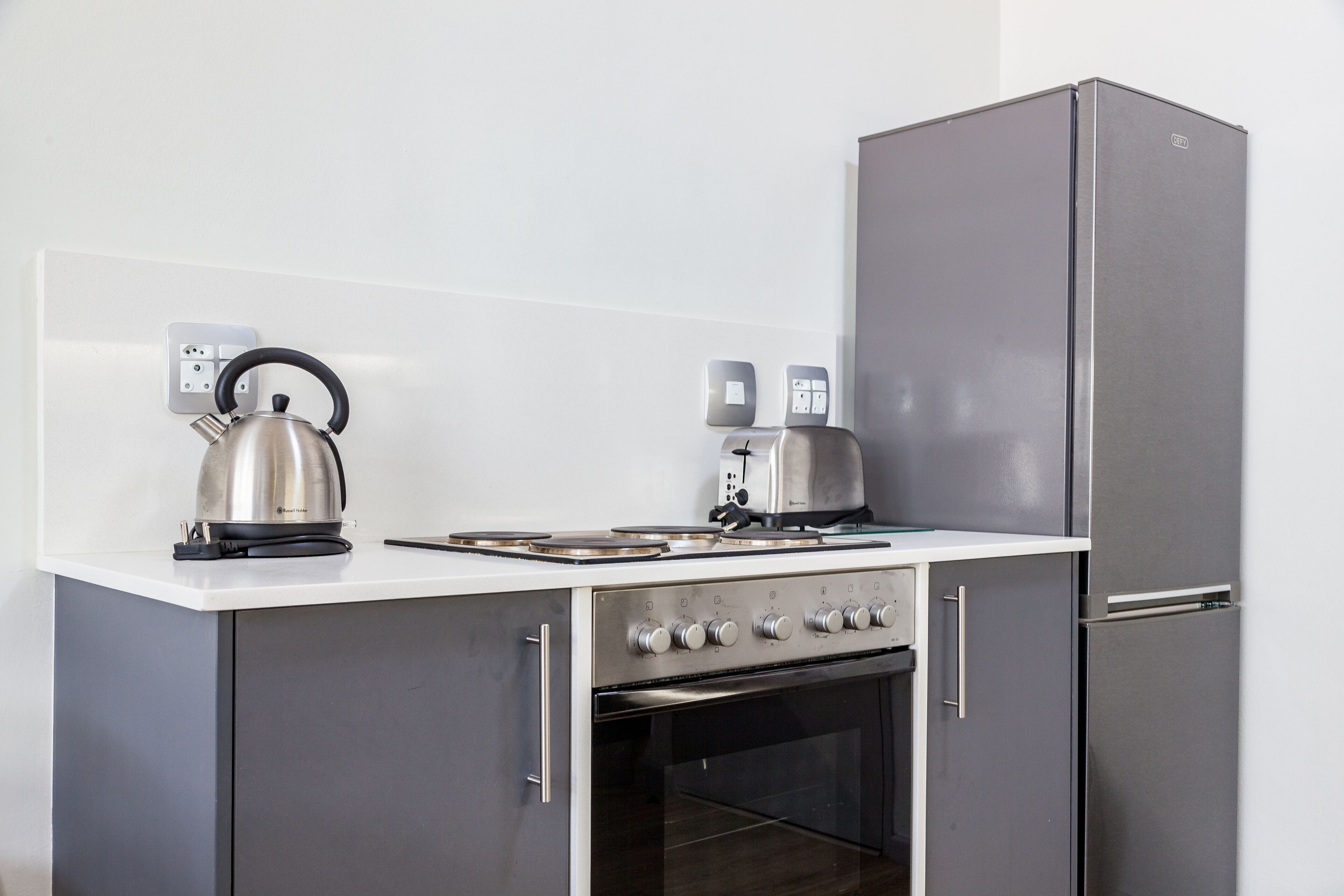 A compact kitchen with a stove, kettle, toaster, and a refrigerator.