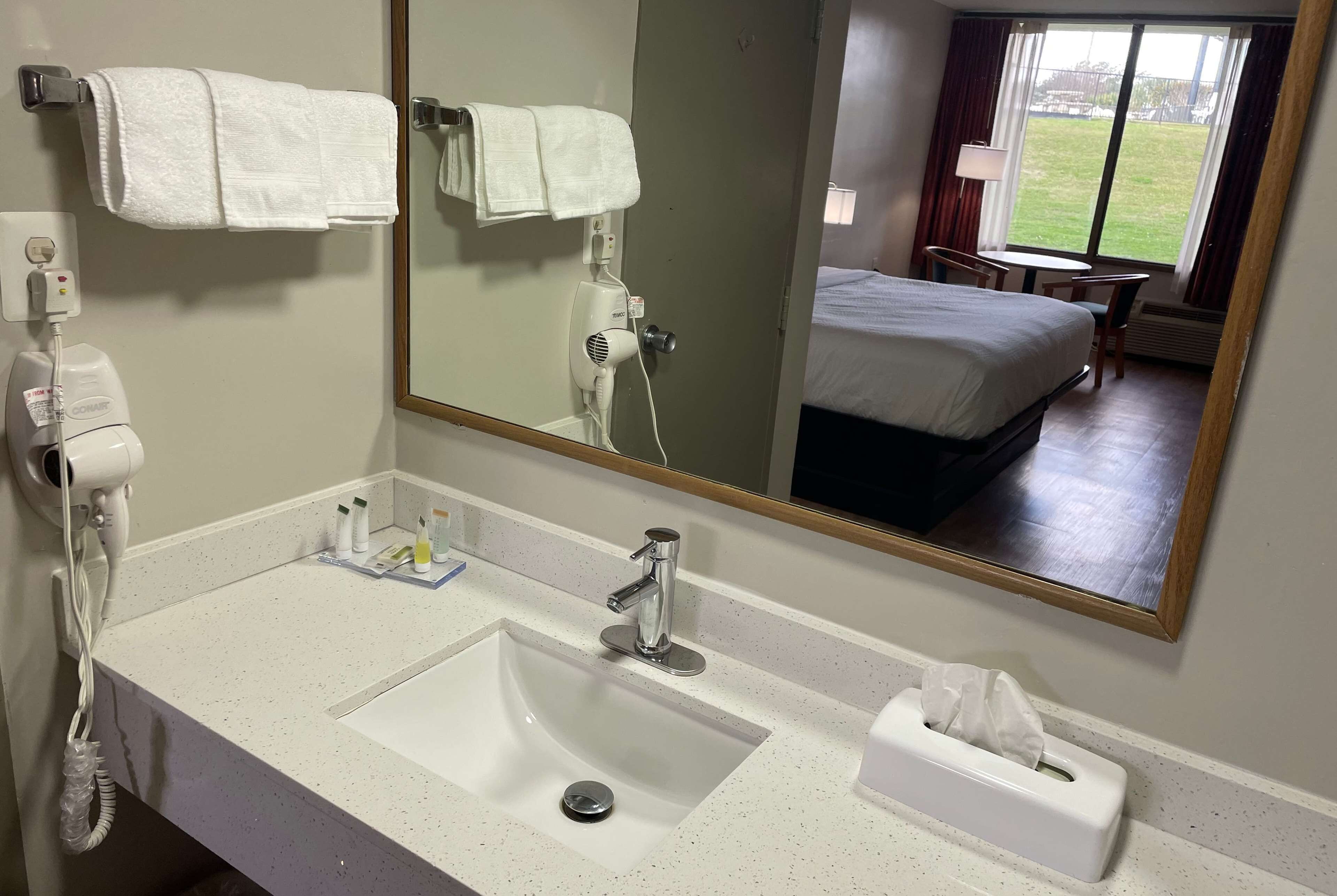 Un baño de hotel con un espejo grande, dos lavamanos, un secador de pelo, un tendedero para toallas y una mesita de noche con lámpara.