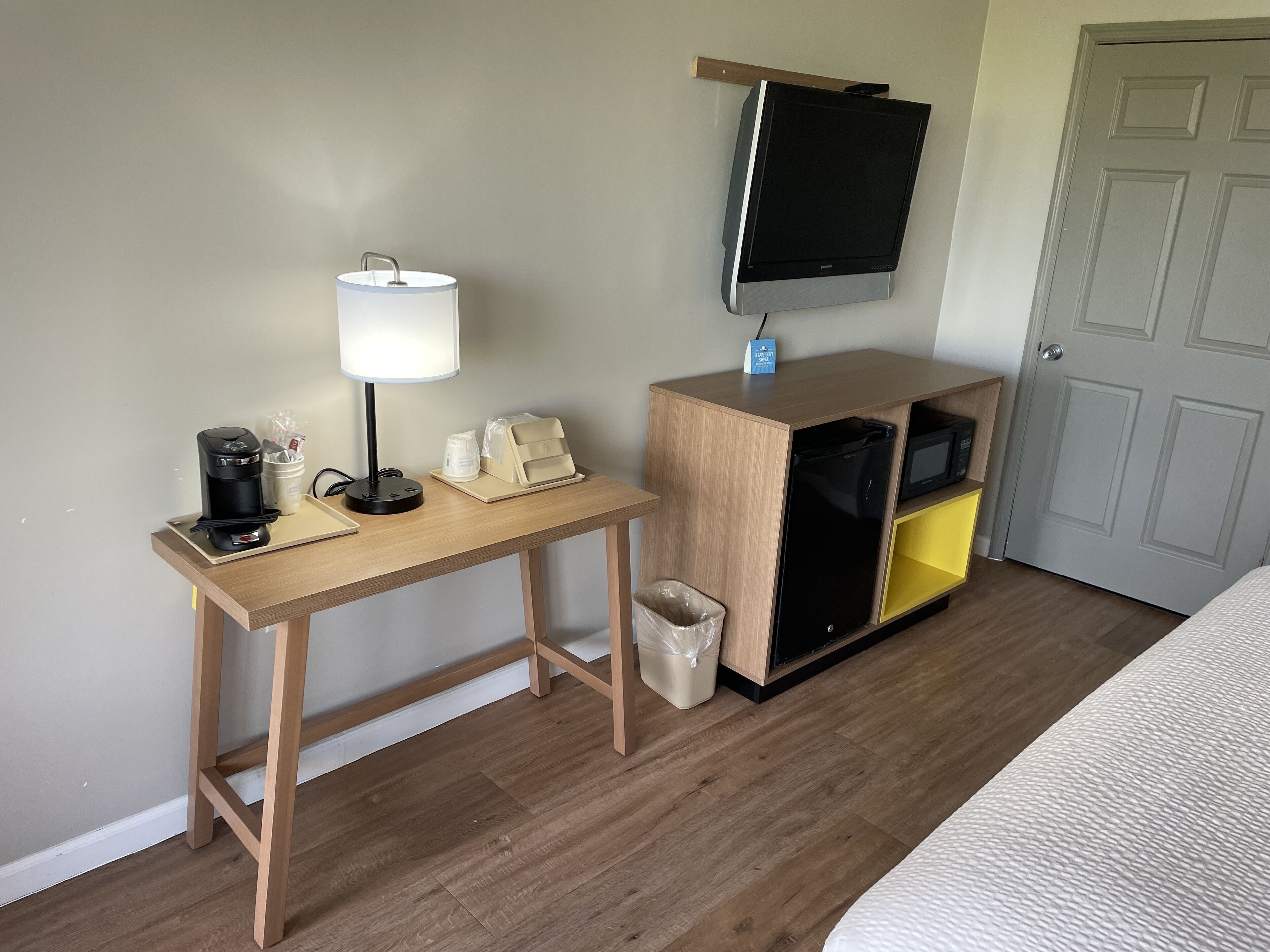 A hotel room with a wooden desk, a coffee maker, a lamp, a trash bin, a TV on a stand, and a bed.