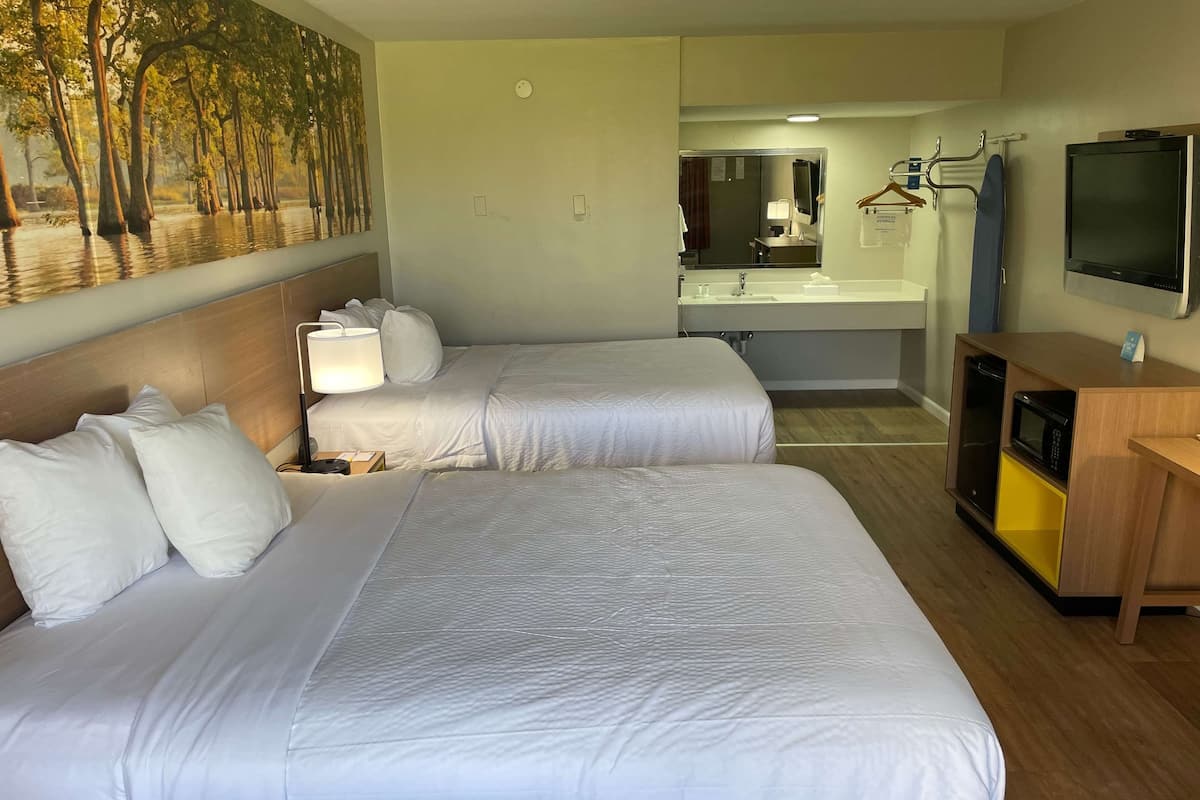 Ein Hotelzimmer mit zwei Betten, einem großen Baumgemälde, einem Badezimmer mit Waschbecken und Spiegel sowie einem Fernseher an der Wand.