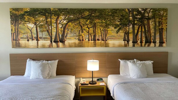 A hotel room with two beds, a nightstand with a lamp, a phone, and a painting of a tree-lined river. - Days Inn by Wyndham Valdosta/Near Valdosta Mall (Valdosta)