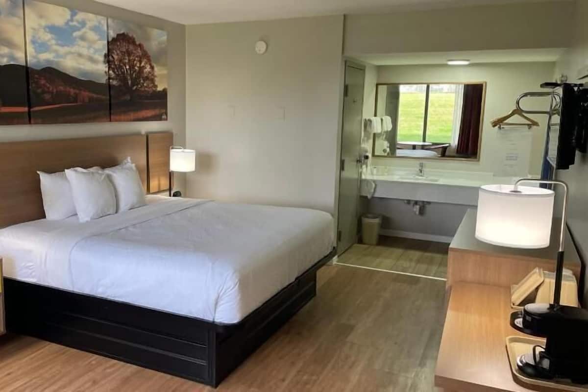 Ein Hotelzimmer mit einem großen Bett, Nachttischlampen, einem Nachttisch, einem Spiegel, einem Badezimmer mit Waschbecken und Spiegel sowie Blick ins Freie.