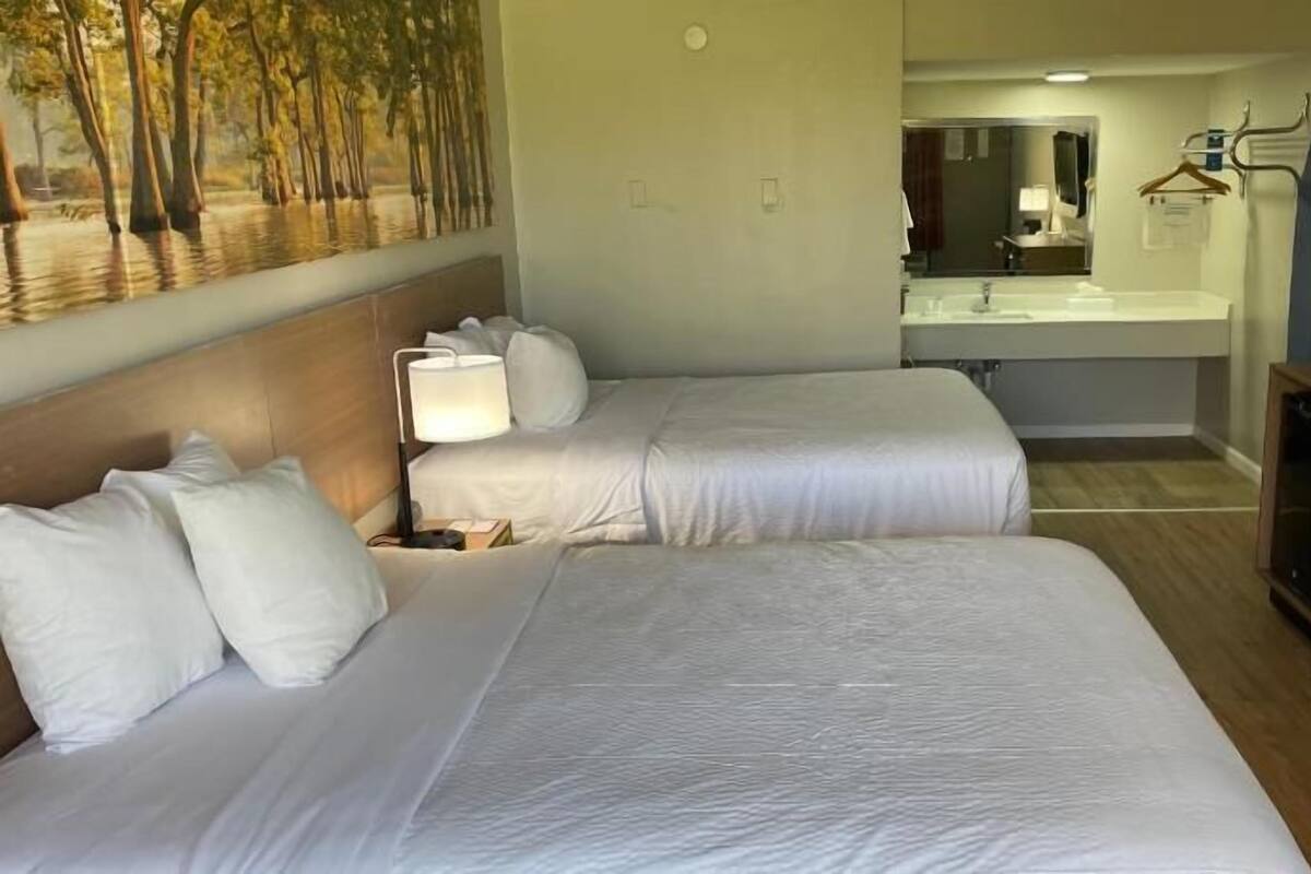 Ein Hotelzimmer mit zwei Betten, einem Badezimmer mit Waschbecken und Spiegel sowie einem Fernseher.