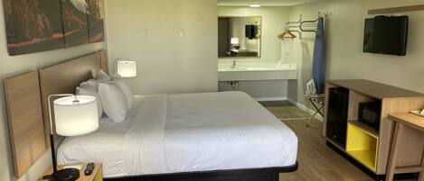A hotel room with a bed, bedside lamp, TV, and a desk with a computer.