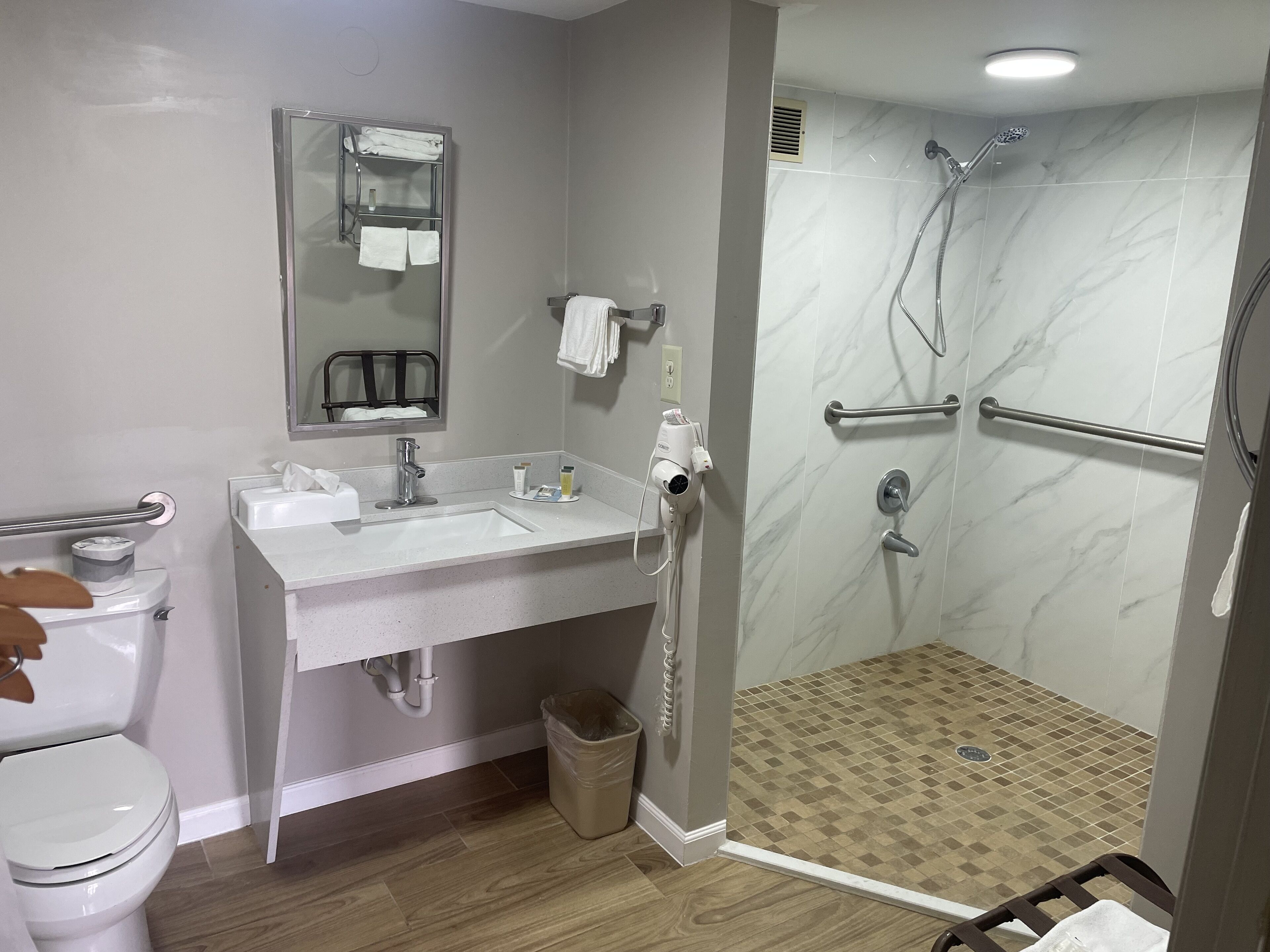 A modern bathroom with a white sink, mirror, and toilet. It includes a shower area with a handheld showerhead and a grab bar.