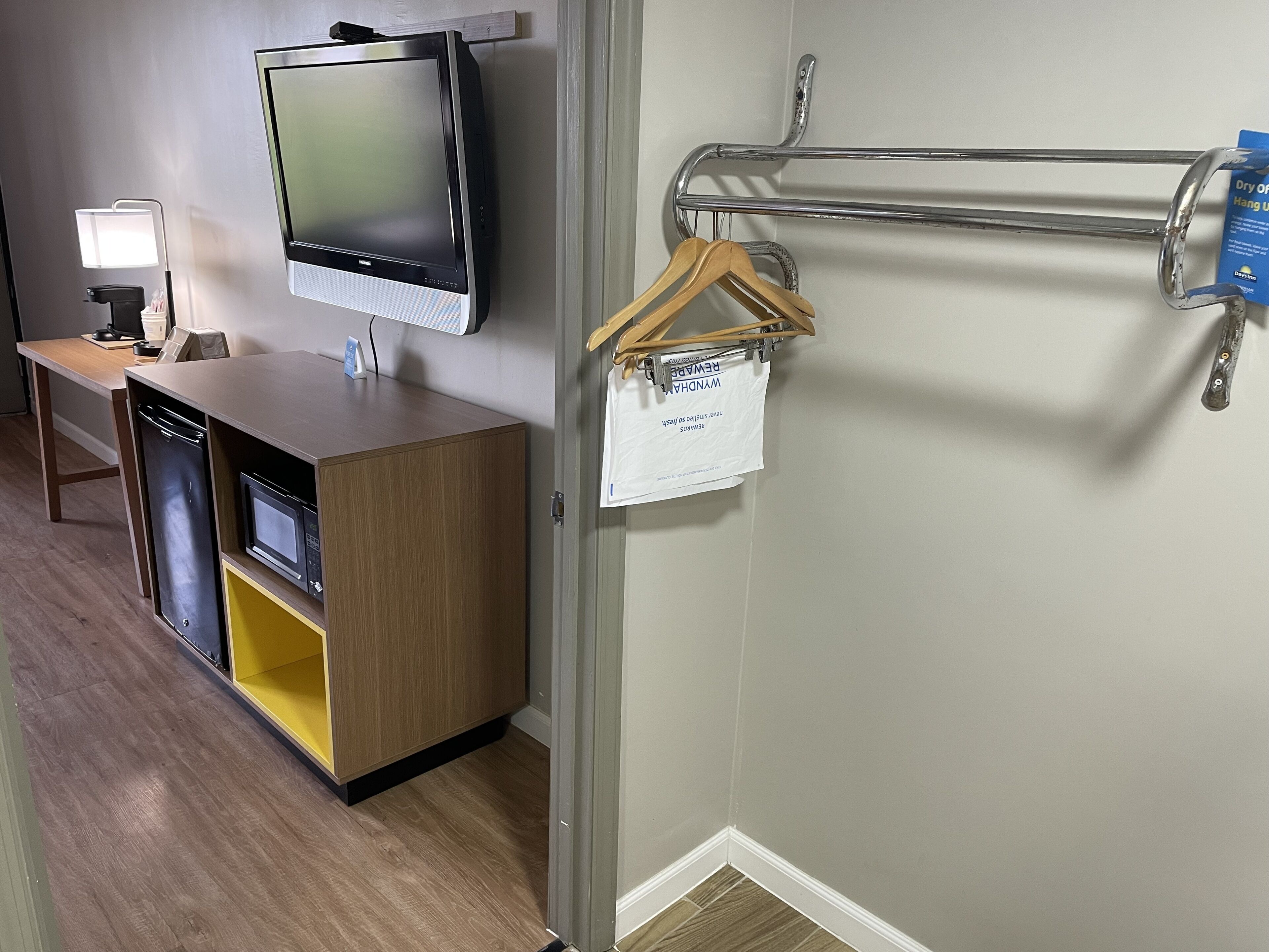 Ein Hotelzimmer mit Flachbildfernseher an der Wand, einem Holzschrank mit Mikrowelle, einem kleinen Tisch mit Lampe sowie einem Metallgestell mit Kleiderbügeln und Handtuch.