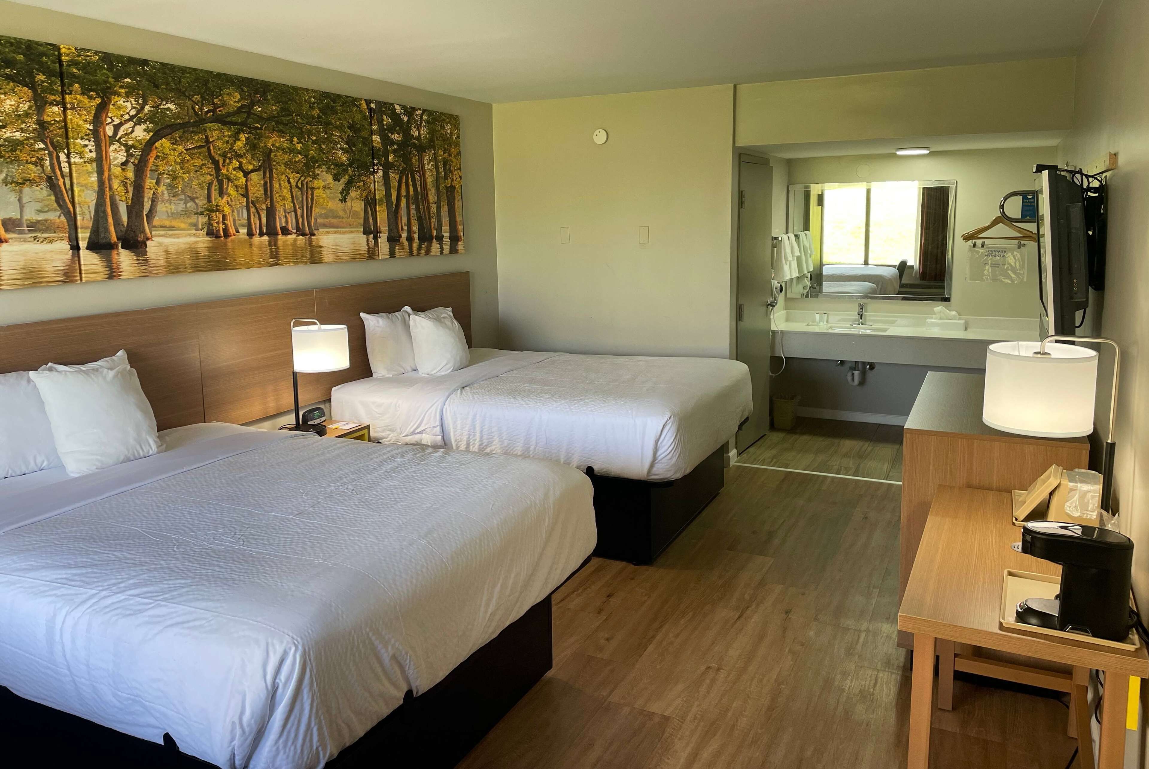 A hotel room with two beds, a large mirror, a small table with a coffee maker, and a painting of trees on the wall.