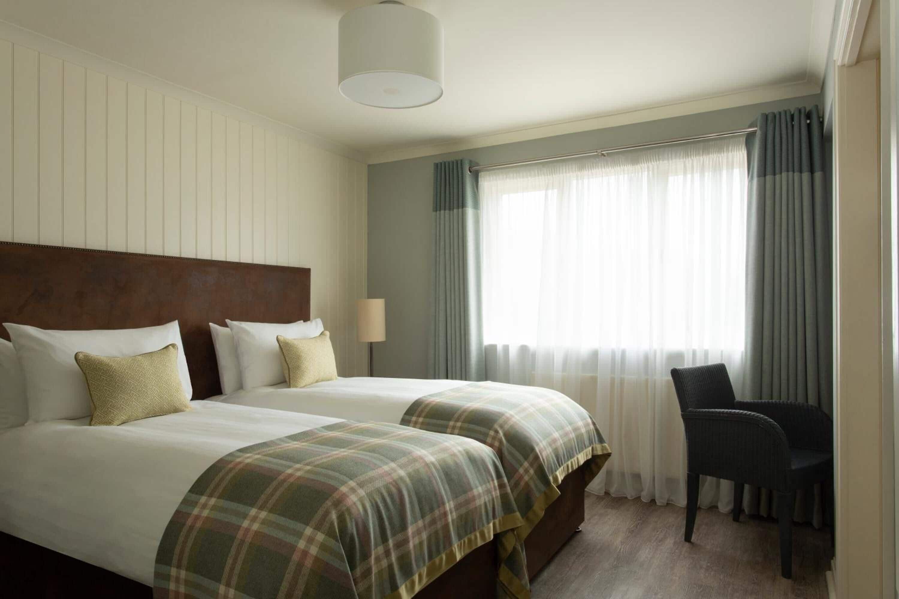 A hotel room with two beds, a headboard, a chair, and a window with curtains.