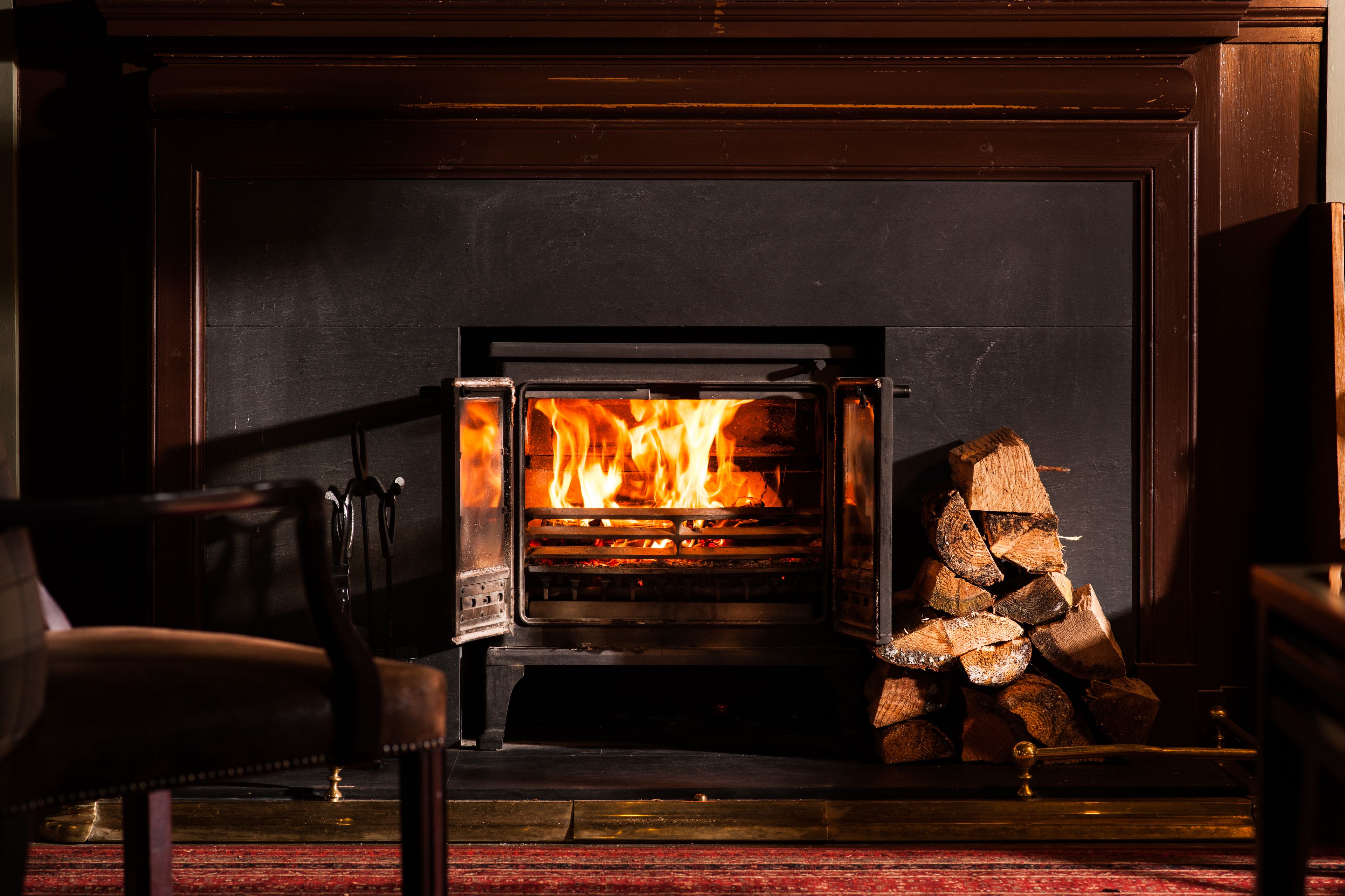 A wood-burning stove with a fire burning inside, a stack of firewood to the right, and a chair in the foreground.