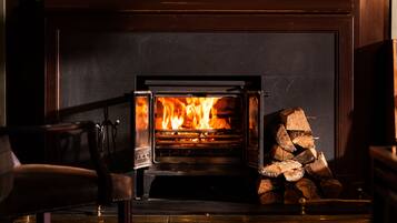 A wood-burning stove with a fire burning inside, a stack of firewood to the right, and a chair in the foreground.