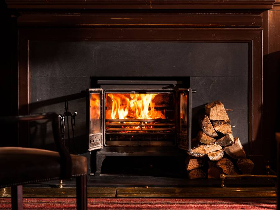 A wood-burning stove with a fire burning inside, a stack of firewood to the right, and a chair in the foreground.