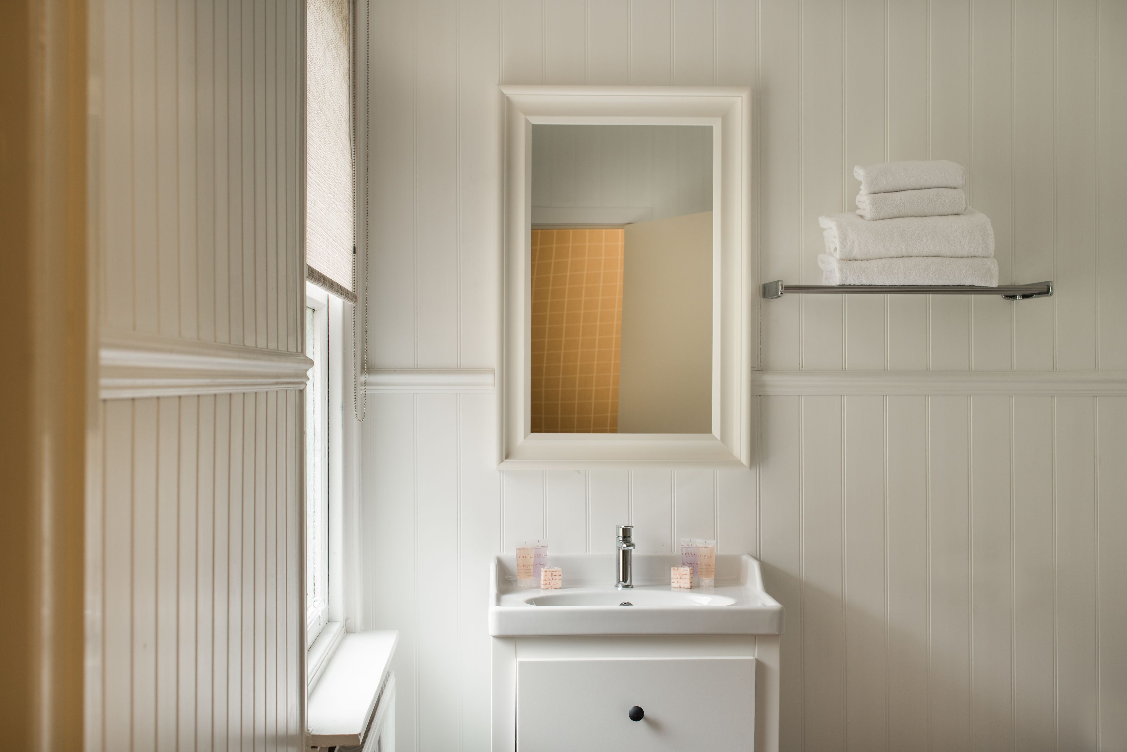 Een badkamer met een witte wastafel, een spiegel, een handdoekhouder en een raam met zonwering.