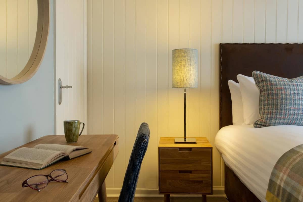 A neatly arranged hotel room with a wooden desk, a lamp, a bed with plaid bedding, and a houndstooth pillow.
