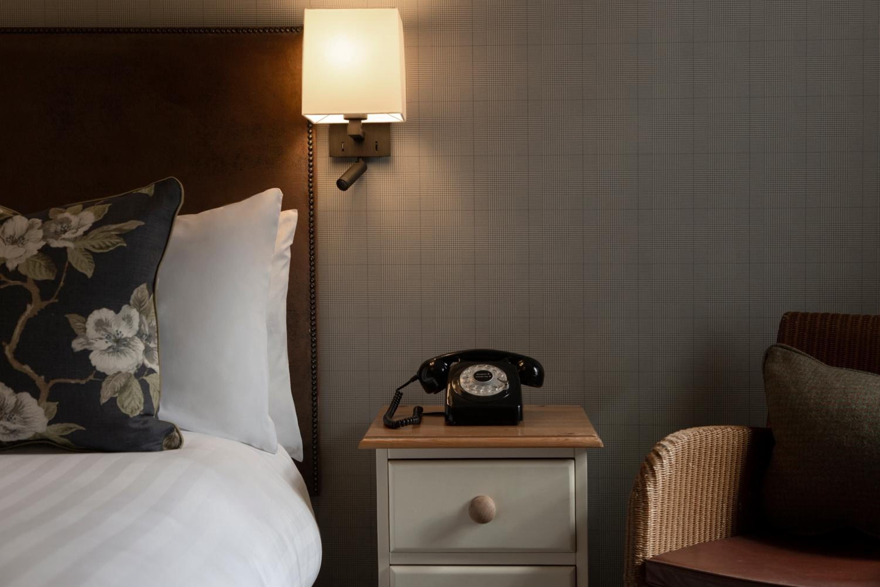 Una habitación de hotel con una cama, una mesita de noche con teléfono, una lámpara y una silla de mimbre.