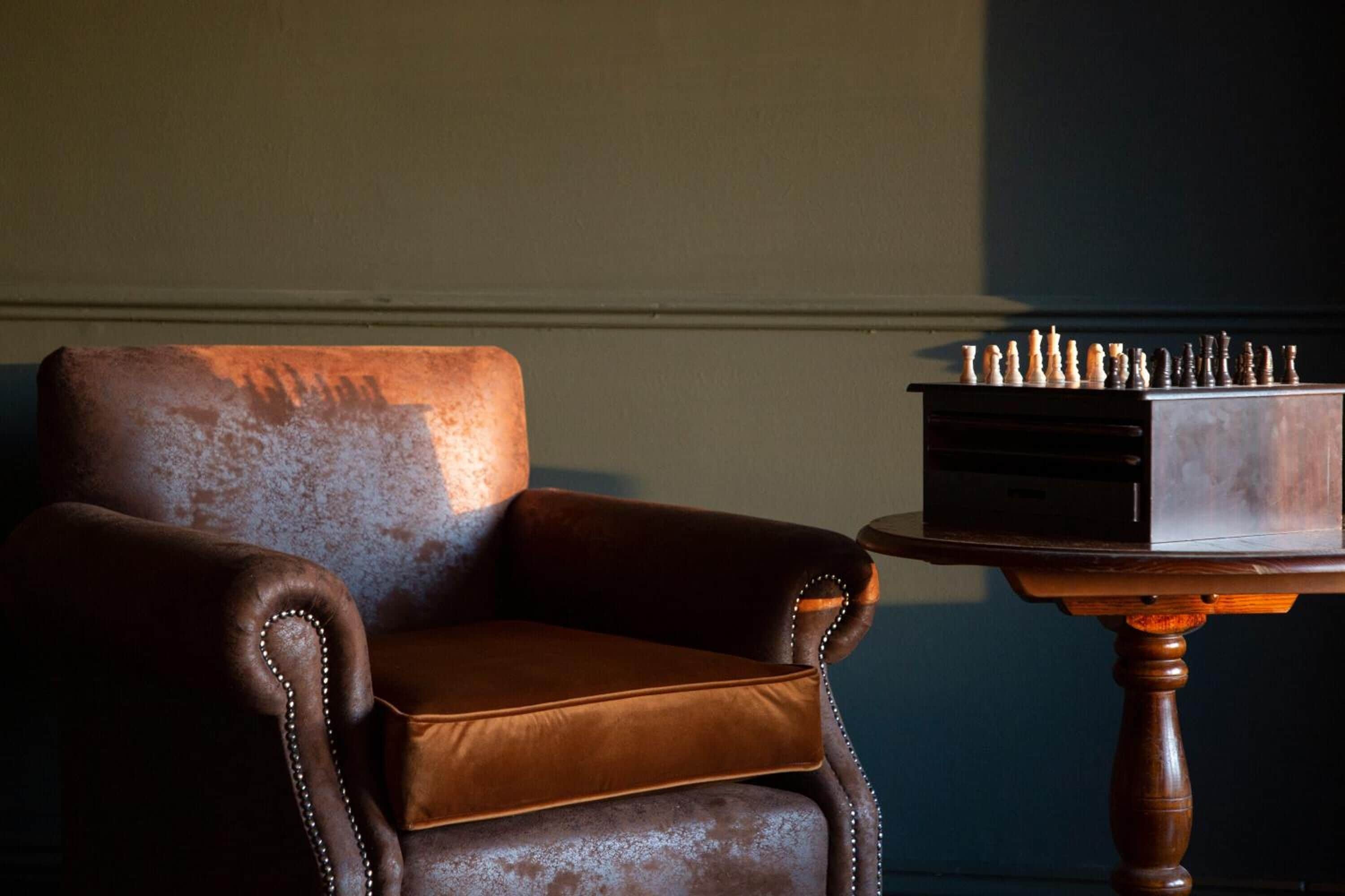 A brown leather armchair with a cushion, a wooden table with a chessboard on it, and a wall in the background.