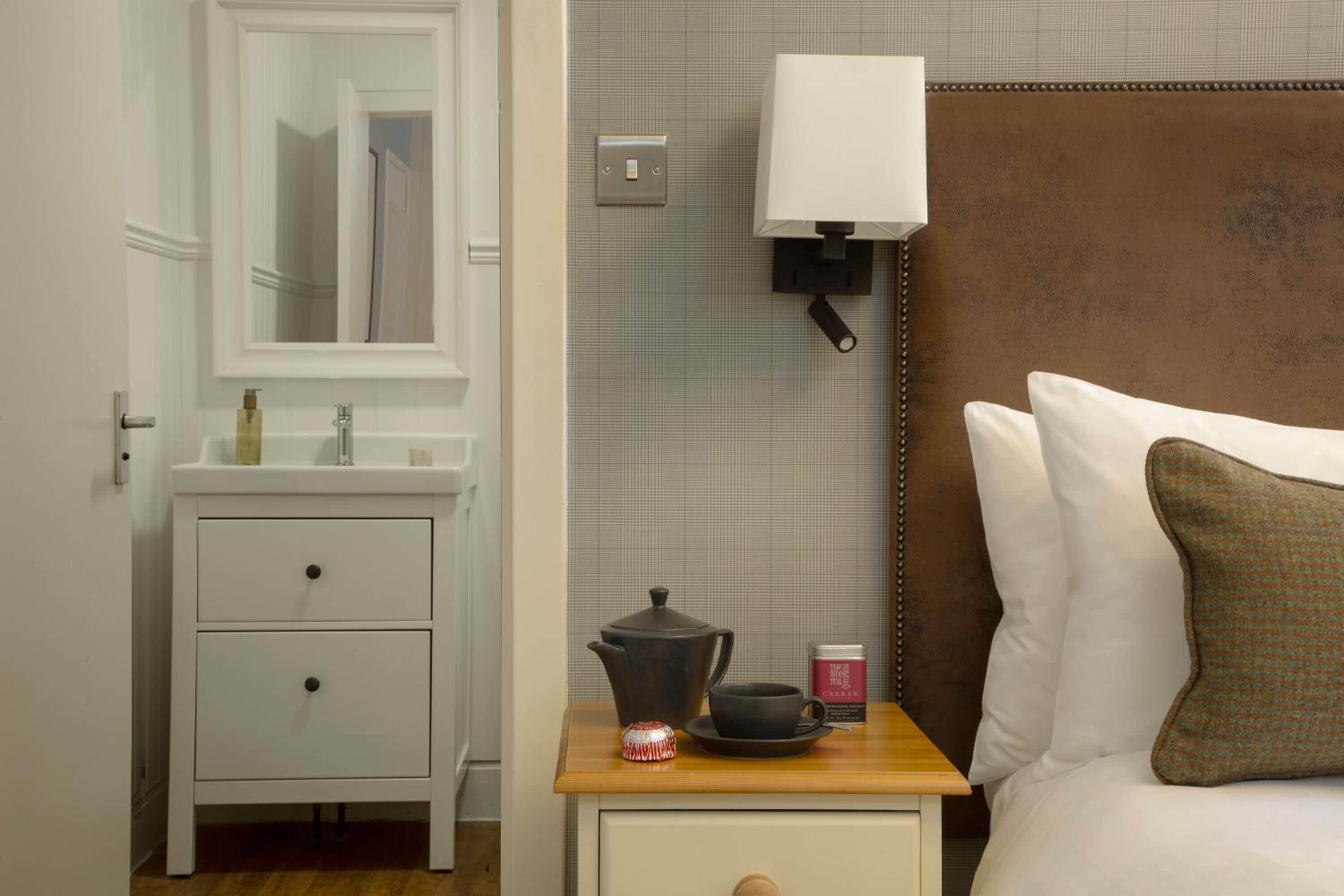 A hotel room with a white dresser, a bedside table with a lamp, a bed with white and brown pillows, and a wall-mounted light switch.