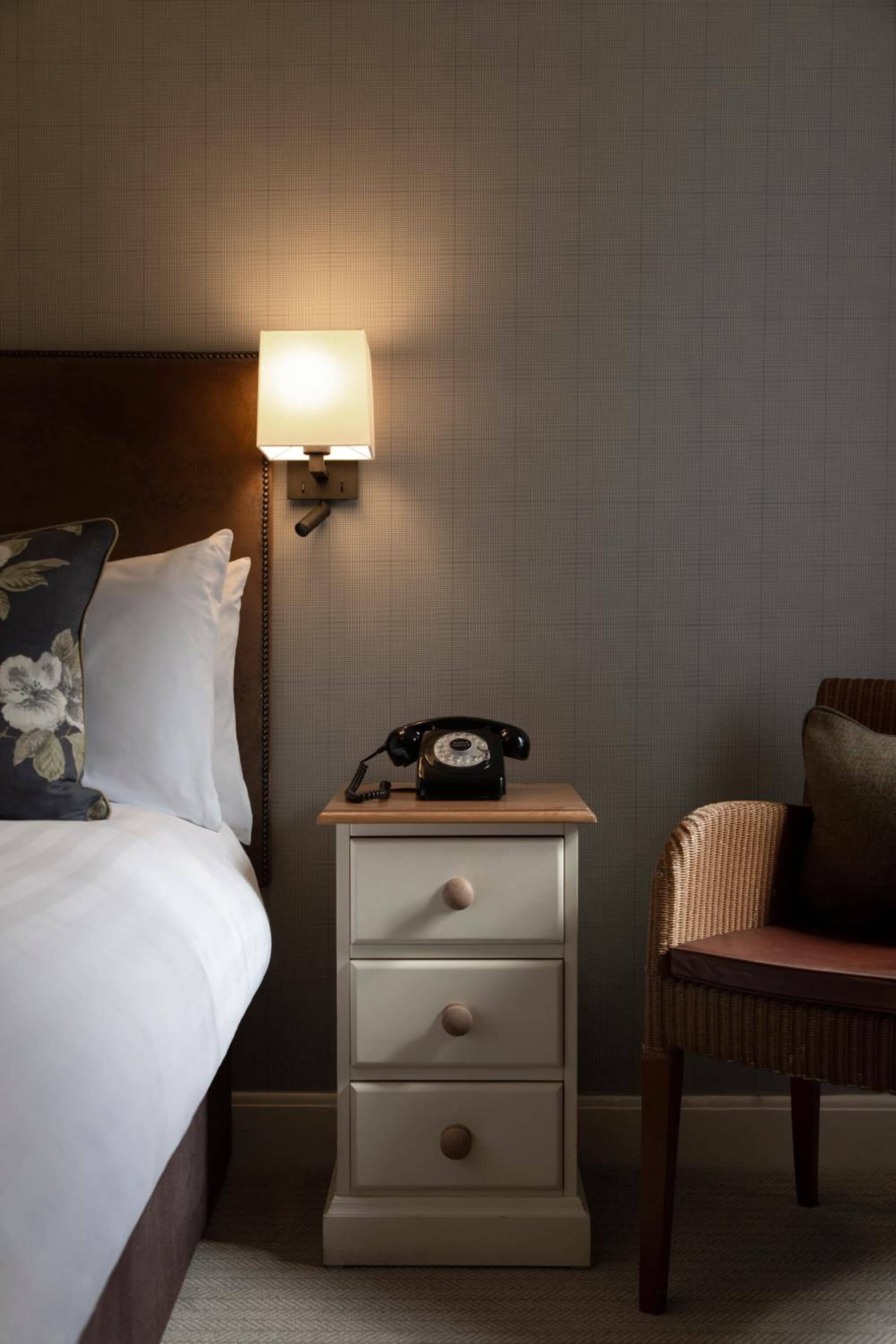 Ein Hotelzimmer mit einem Bett, einem Nachttisch mit Lampe, einem Telefon und einem Korbstuhl.