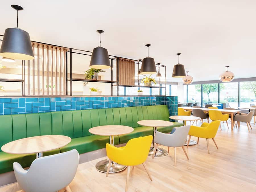 Ein modernes Café-Interieur mit grünen Sitzgelegenheiten, gelben Stühlen und mehreren Tischen.