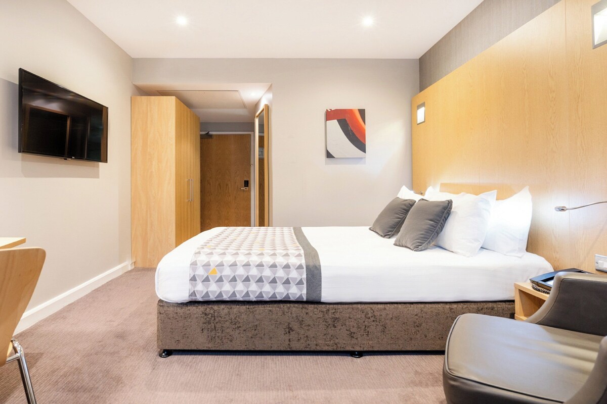 A modern hotel room with a bed, a desk, a chair, a TV, and a door leading to another room.