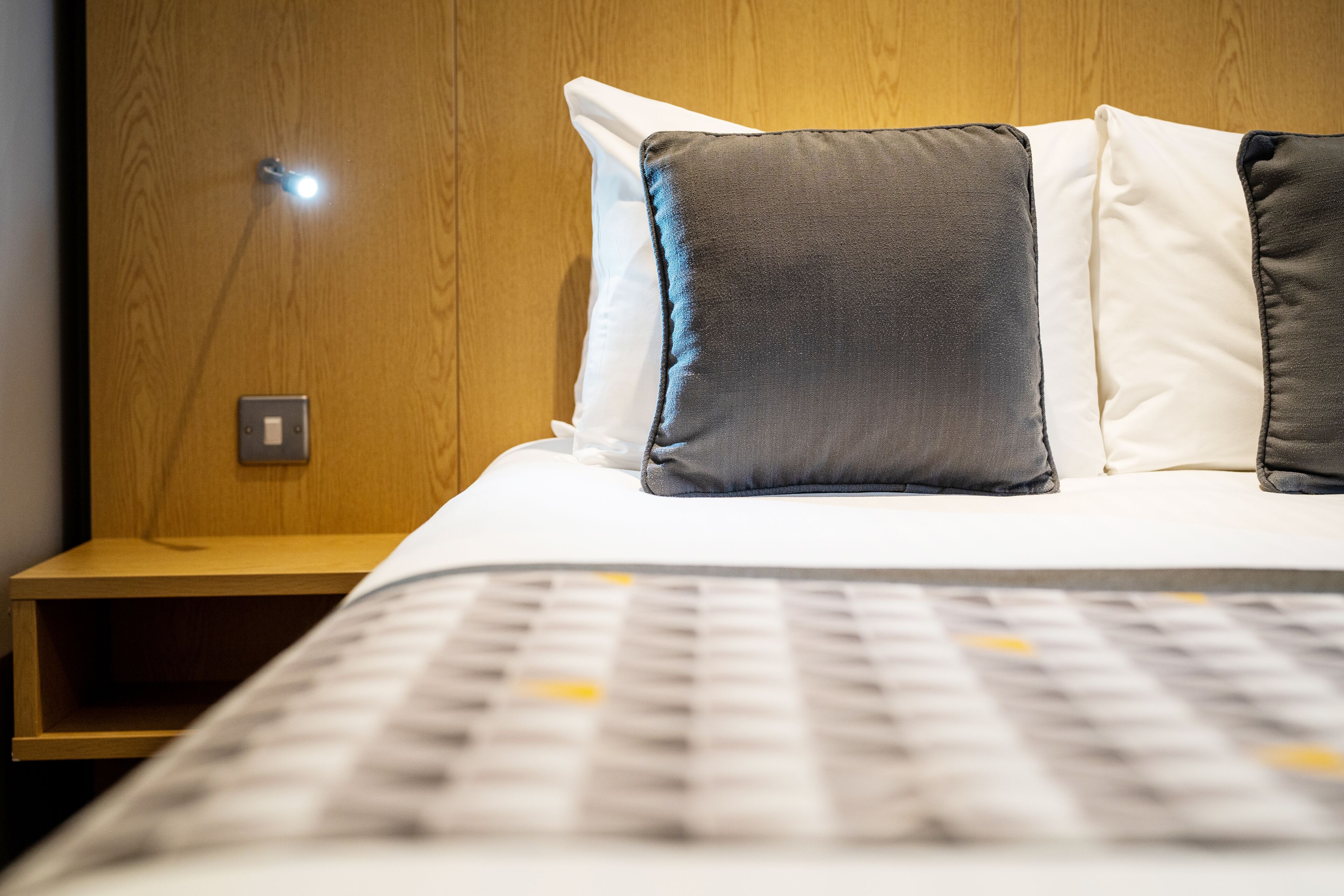 A neatly made bed with a patterned bedspread and two pillows, a wooden nightstand with a light, and a wooden wall.