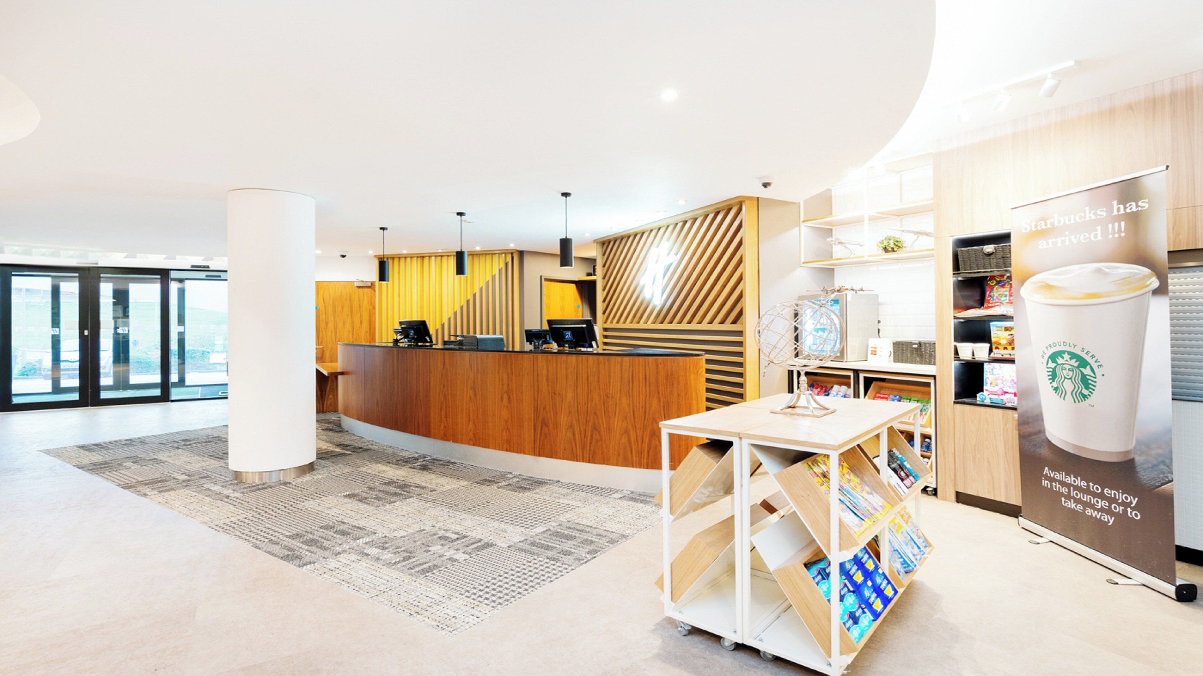A modern hotel lobby with a reception desk, a coffee station, and a view of the outdoors.