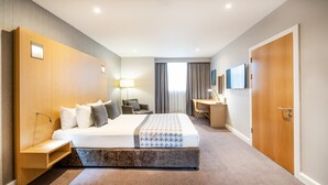 Ett hotellrum med en stor säng, ett skrivbord, en stol, en lampa och en garderob.