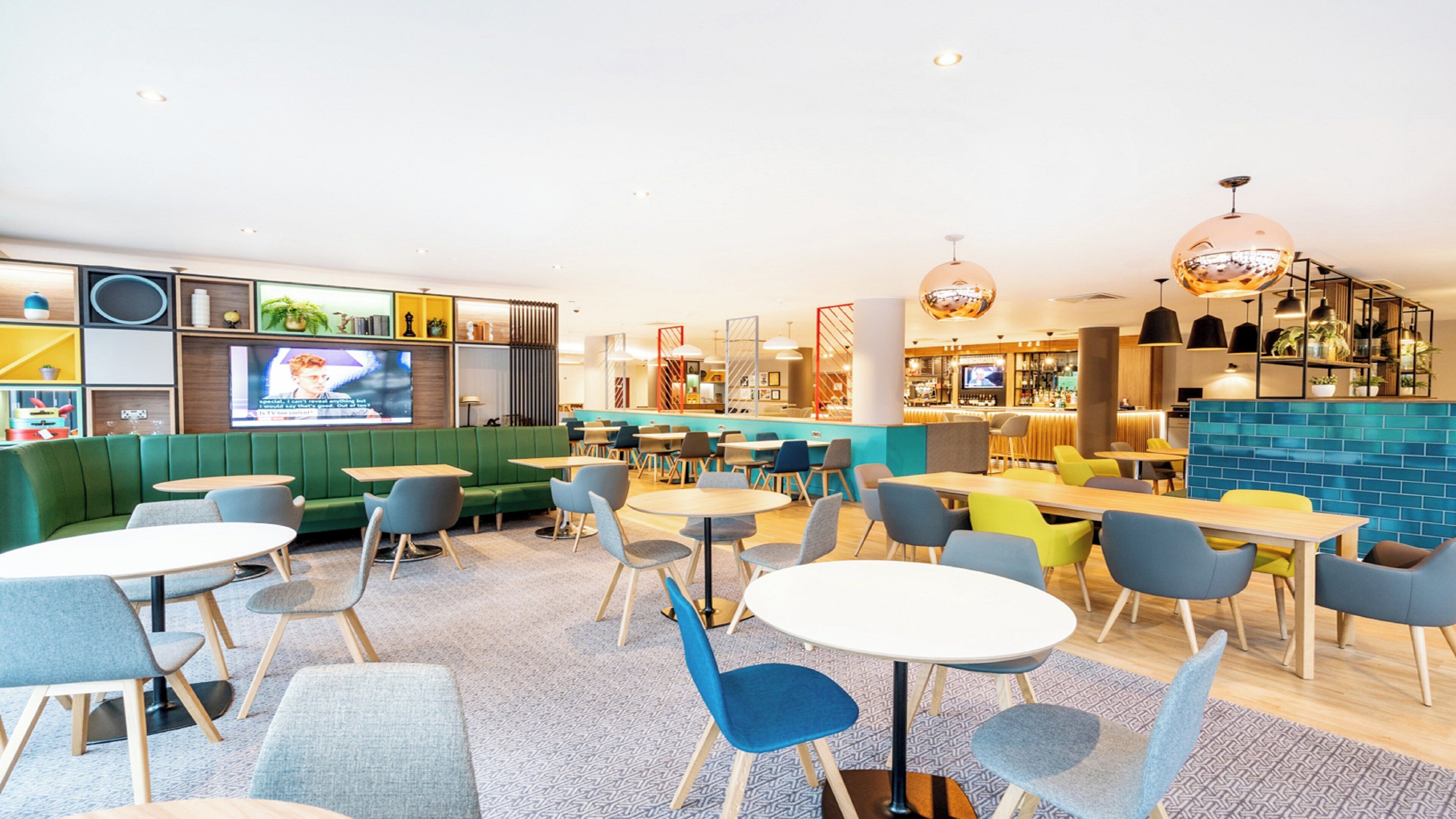 A modern lounge area with green seating, round tables, and a bar in the background.